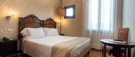 A neatly made bed with a headboard, a bedside table with a lamp, a small round table with a chair, and a window with curtains.