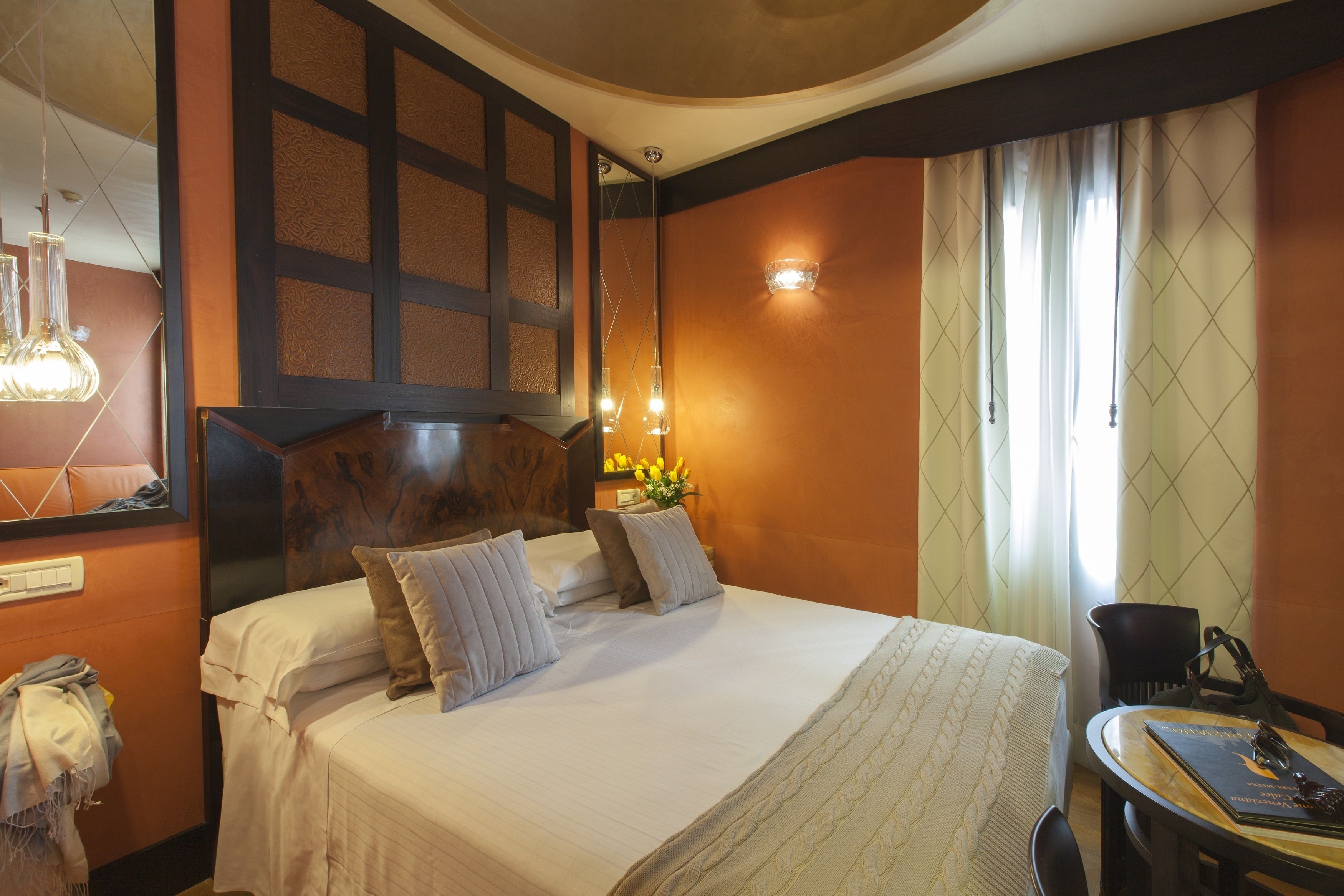 A hotel room with a large bed, a bedside table, a chair, and a small table with a book and a lamp.