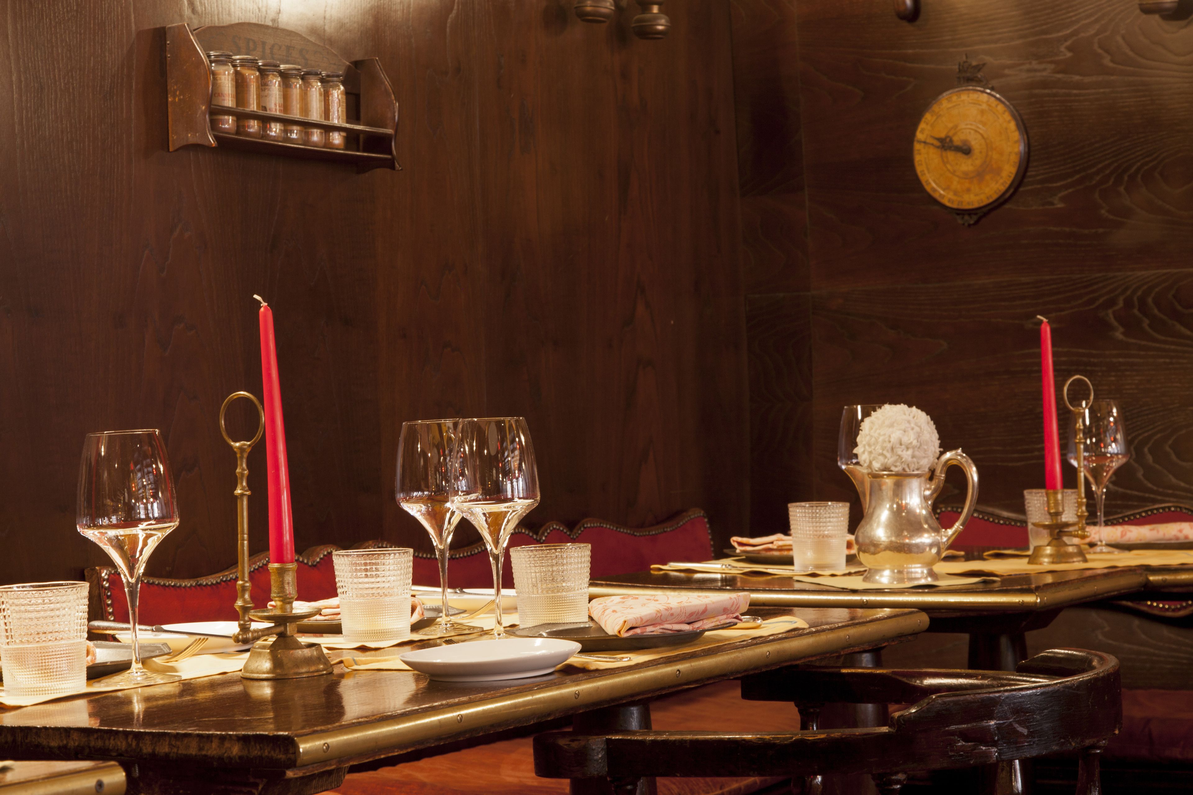 Una mesa de comedor dispuesta con copas de vino, platos y velas, con un muro de madera y un reloj de fondo.