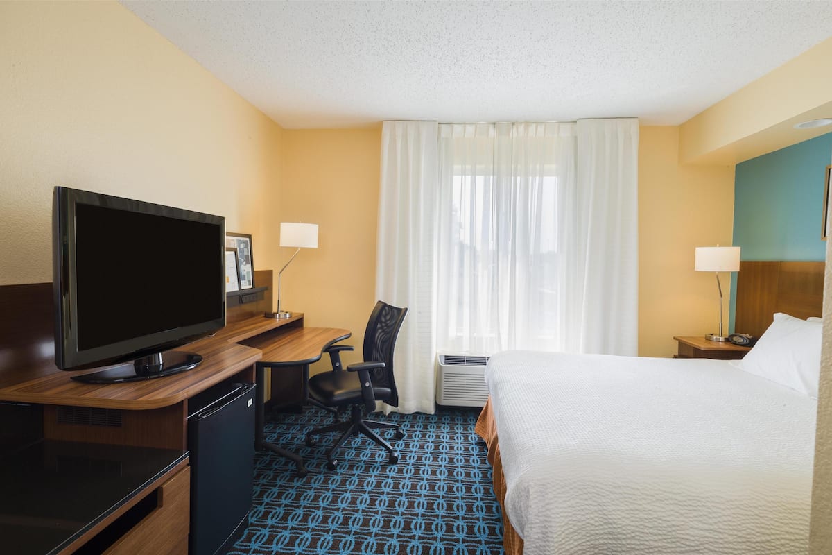 Habitación de hotel con una cama, un escritorio con televisor, una silla, una lámpara de escritorio y una ventana con cortinas.