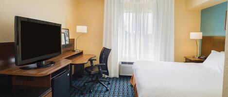 Habitación de hotel con una cama, un escritorio con televisor, una silla, una lámpara de escritorio y una ventana con cortinas.