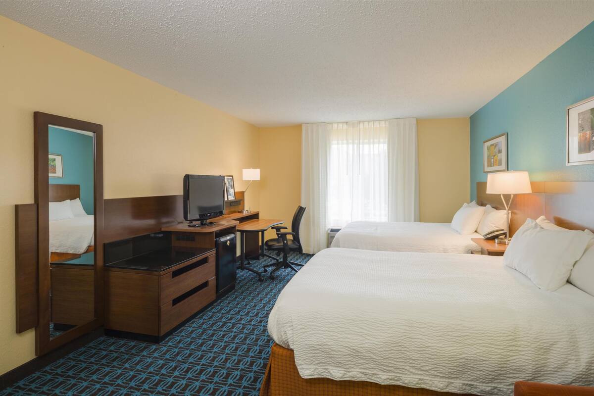Habitación de hotel con dos camas, un escritorio con televisor, un espejo y una ventana con cortinas.