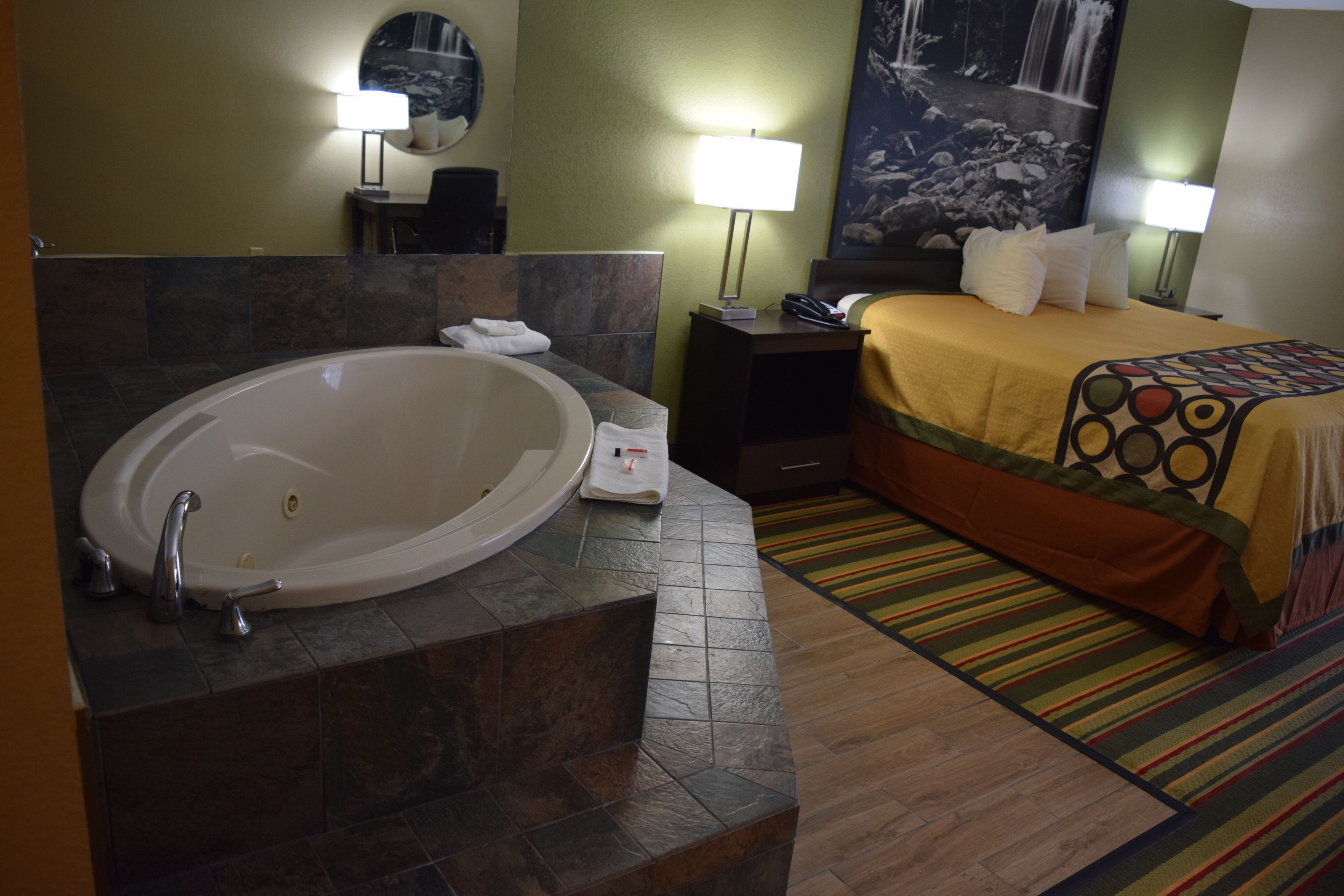 Una camera d'albergo con una grande vasca da bagno, un letto con una coperta colorata, un comodino con una lampada e un quadro appeso al muro.