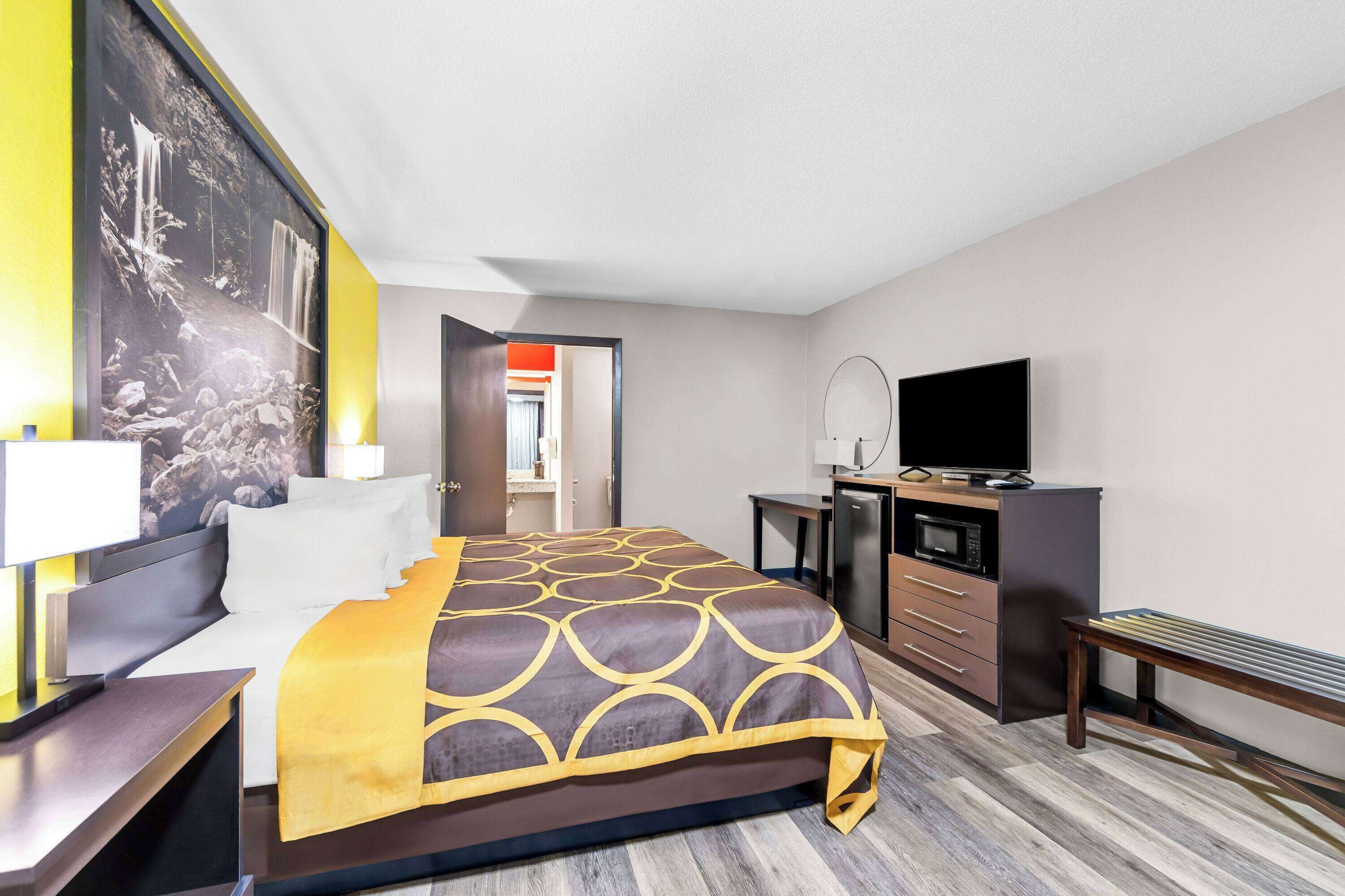 A hotel room with a bed, bedside table, lamp, TV, and a kitchenette area.