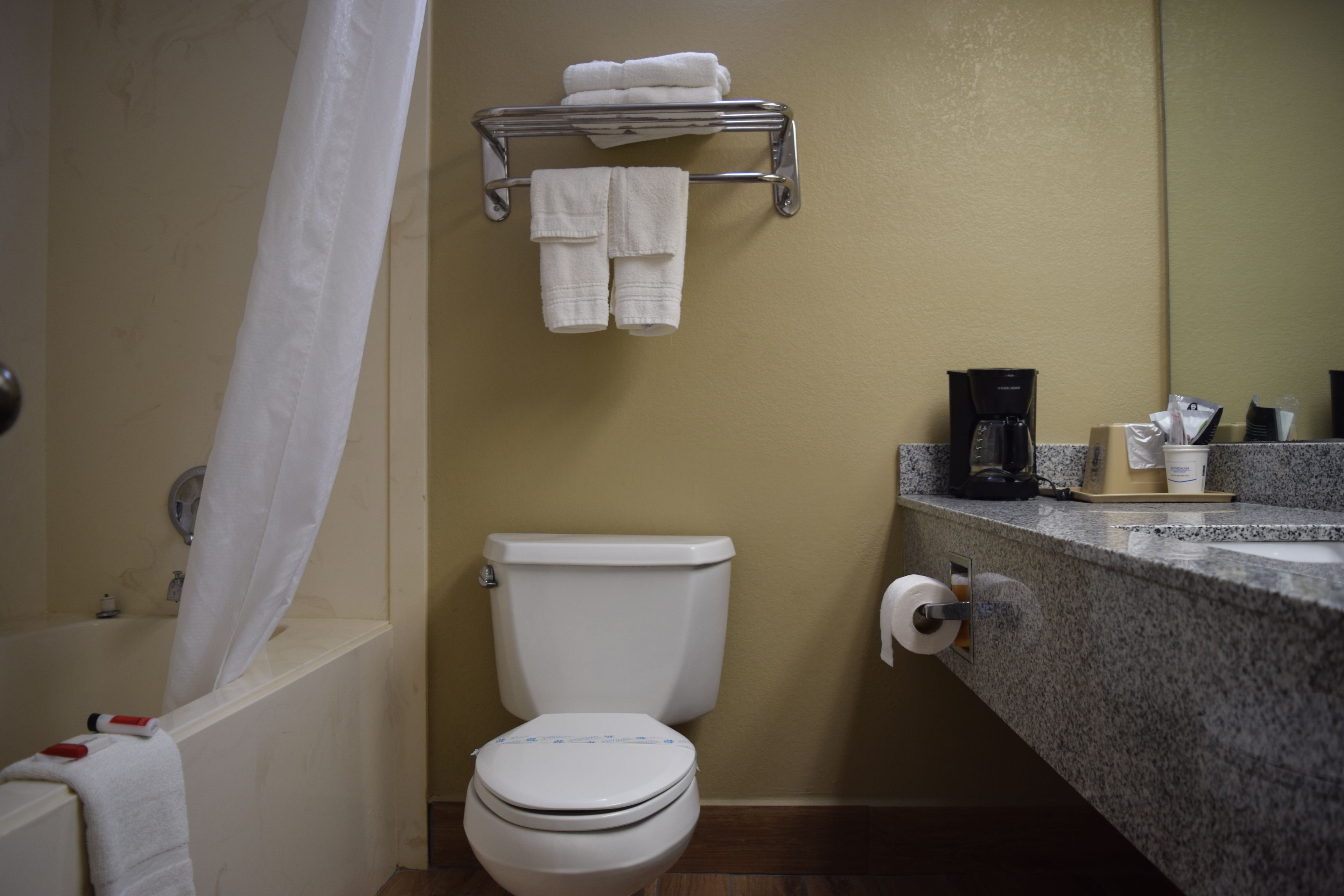 Casa de banho com sanita, um porta-toalhas com toalhas dobradas, máquina de café e dispensador de papel.