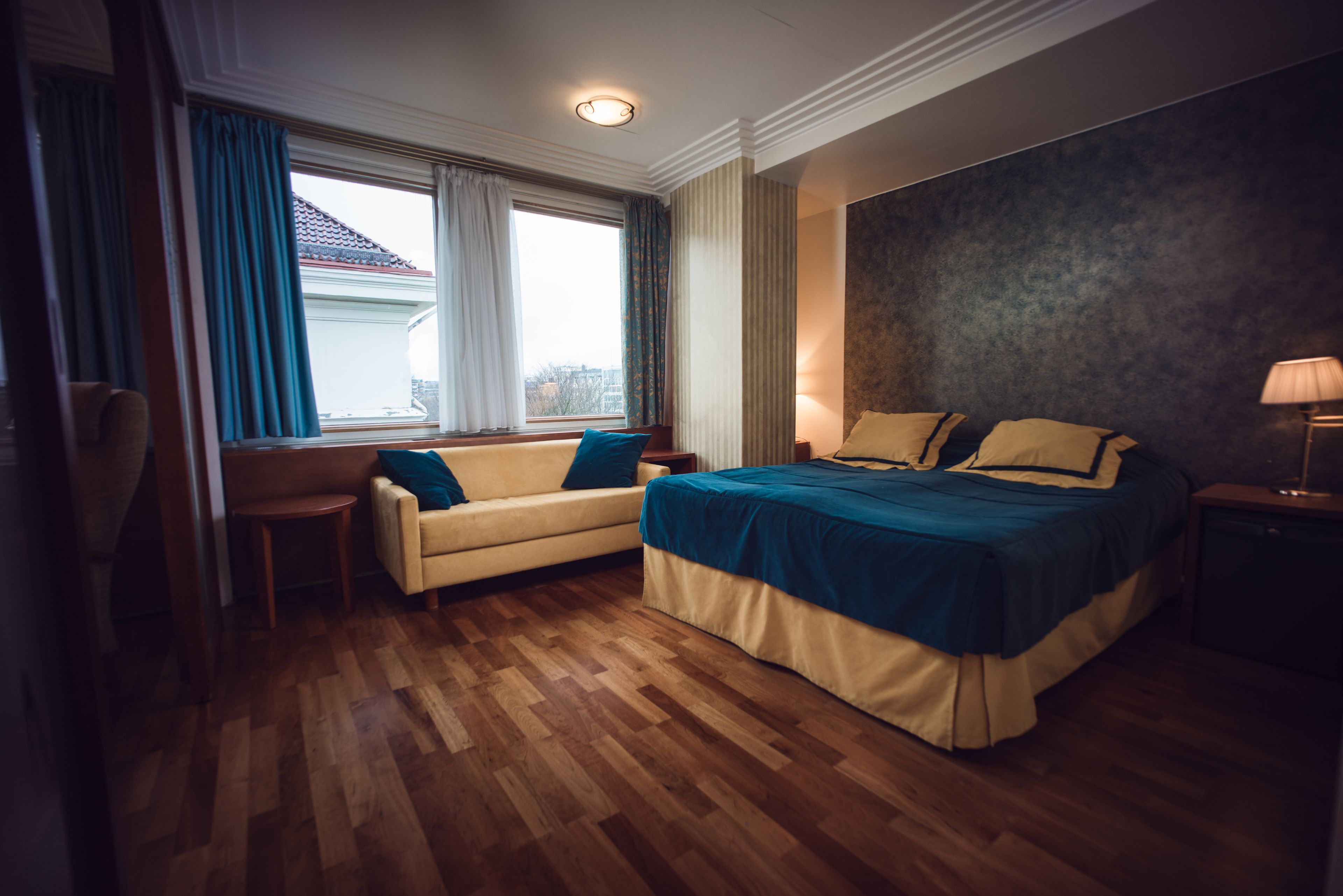 A hotel room with a bed, a sofa, a small table, a lamp, and a window with curtains.