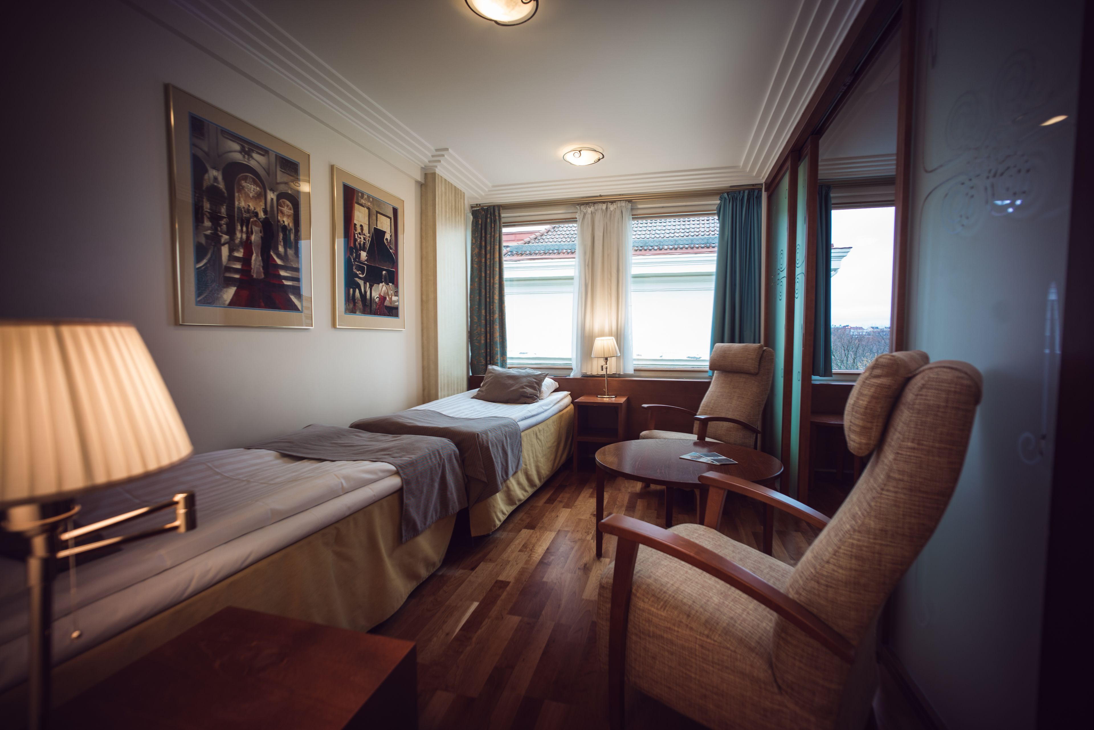 A compact hotel room with two single beds, a small table, a chair, and a window with curtains.