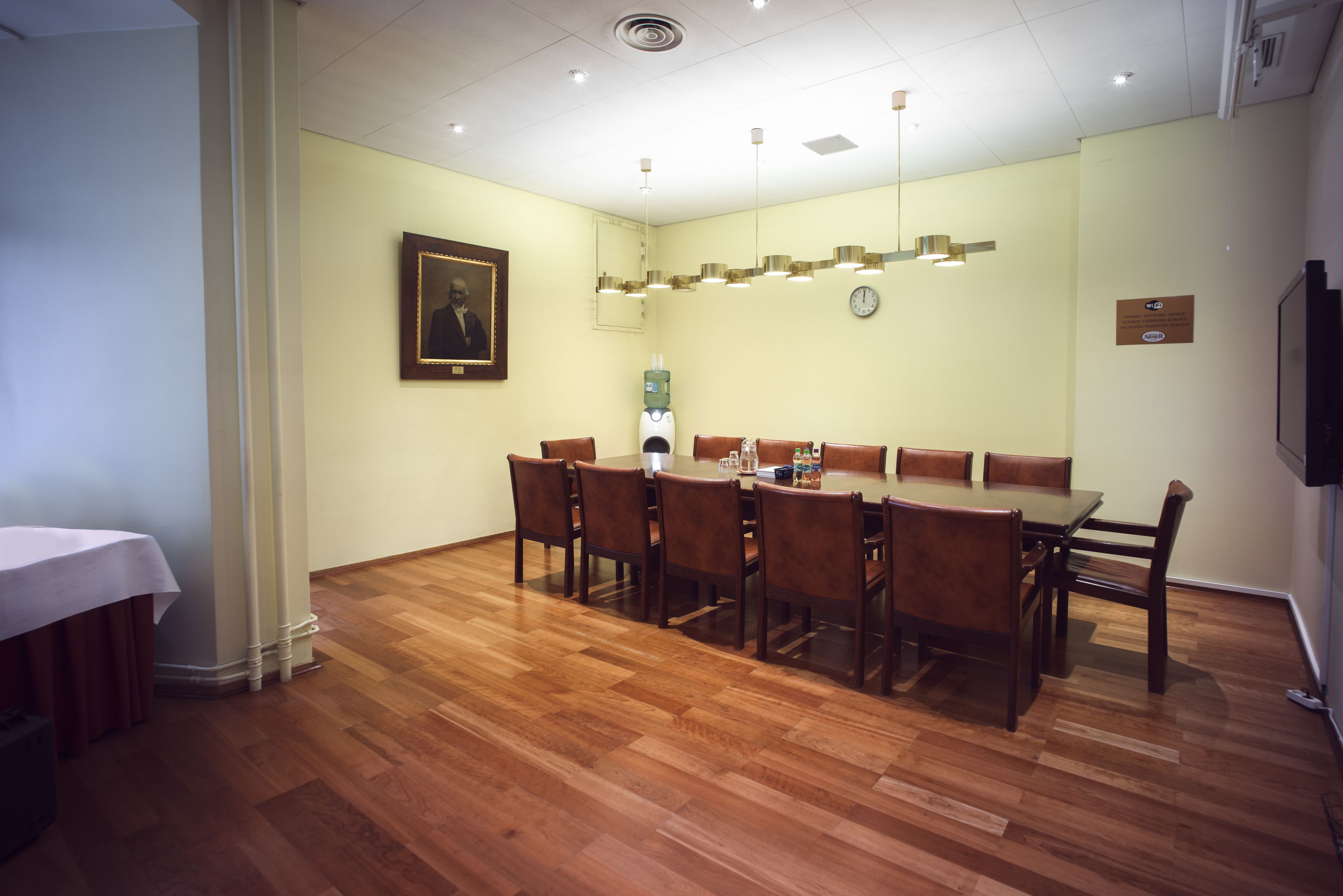 Un salón de conferencias con una mesa y sillas de madera, un dispensador de agua y un reloj en la pared.