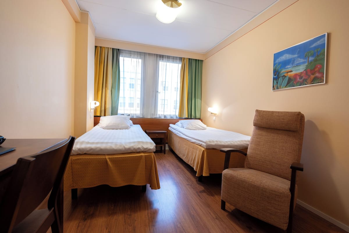 Hotellihuone, jossa on kaksi erillistä sänkyä, puinen työpöytä, tuoli, ikkuna verhoilla ja taulu seinällä.