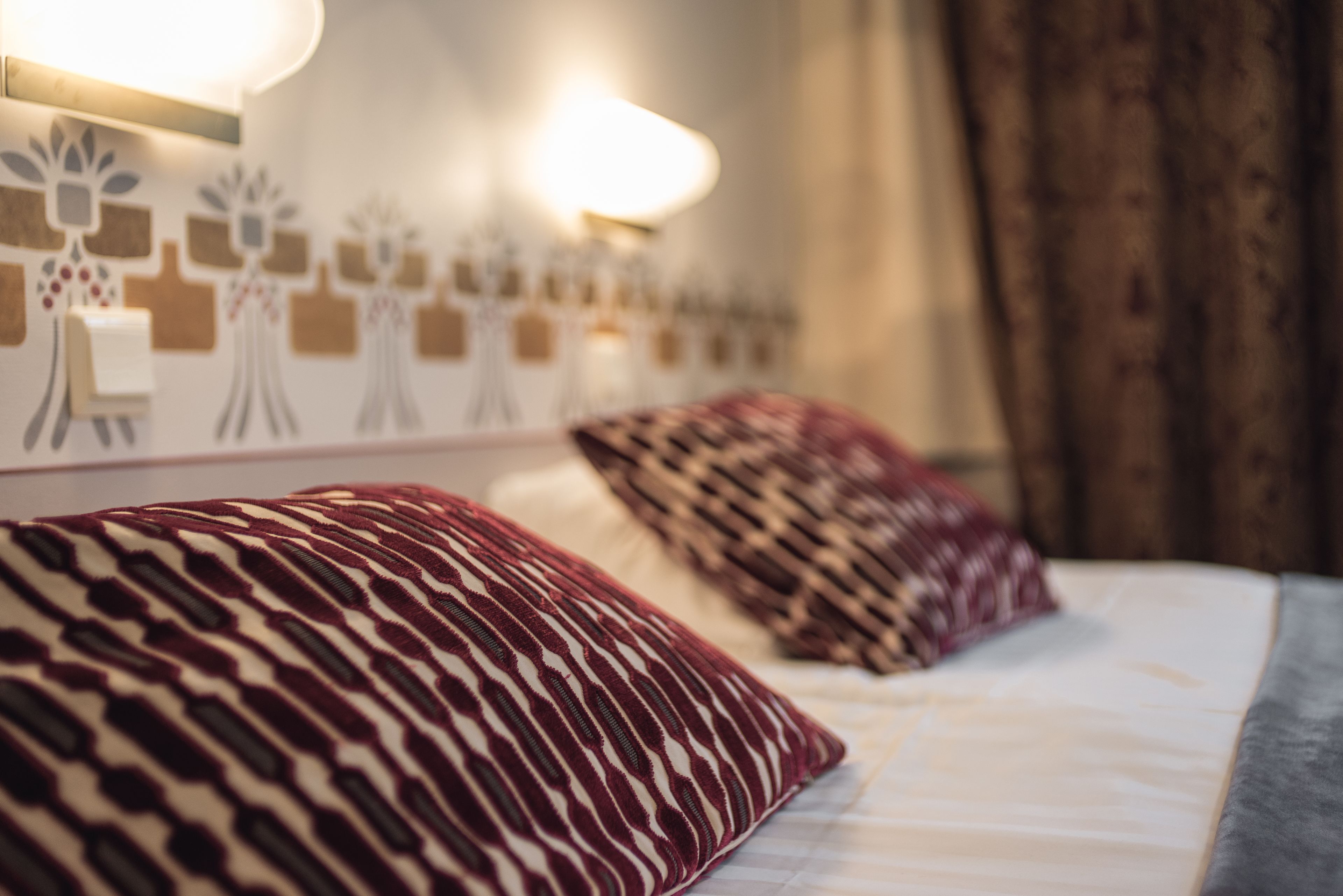 A hotel room with a bed, patterned pillows, a wall-mounted light fixture, and a curtain.
