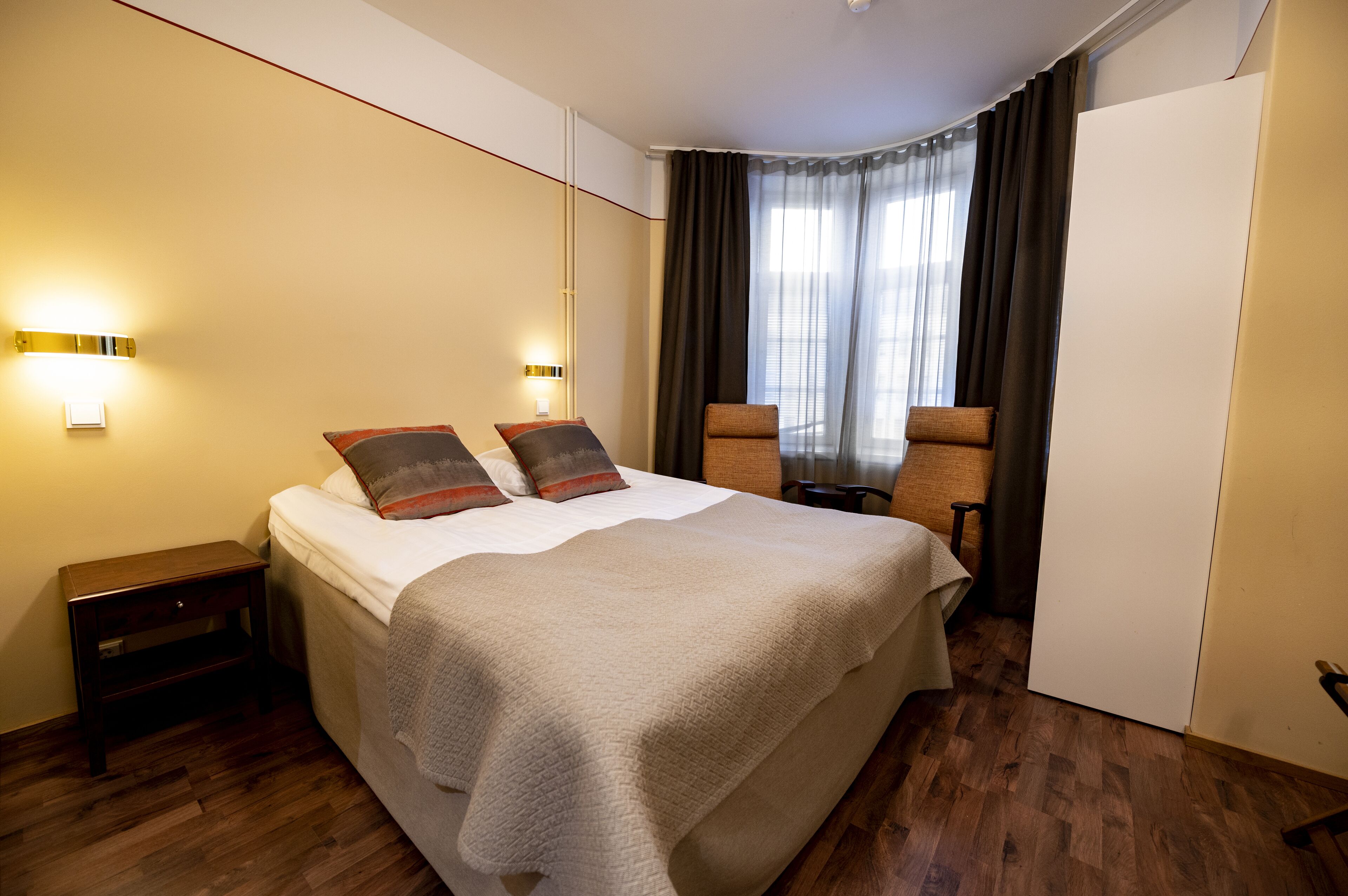 A hotel room with a large bed, two bedside tables, a wardrobe, and a window with curtains.