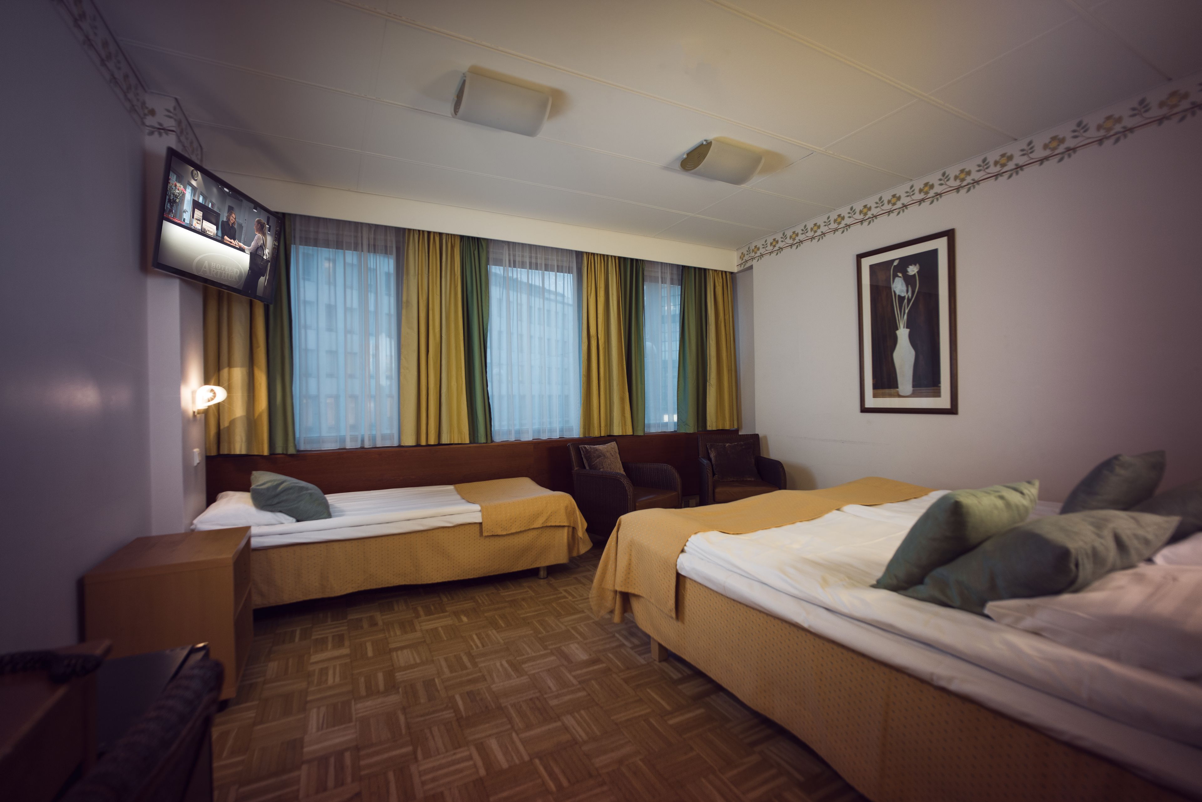 A hotel room with two single beds, a sofa bed, a TV mounted on the wall, and a window with curtains.