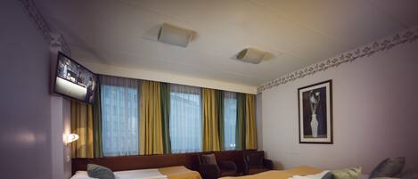 Habitación de hotel con dos camas individuales, un sofá cama, un televisor montado en la pared y una ventana con cortinas.