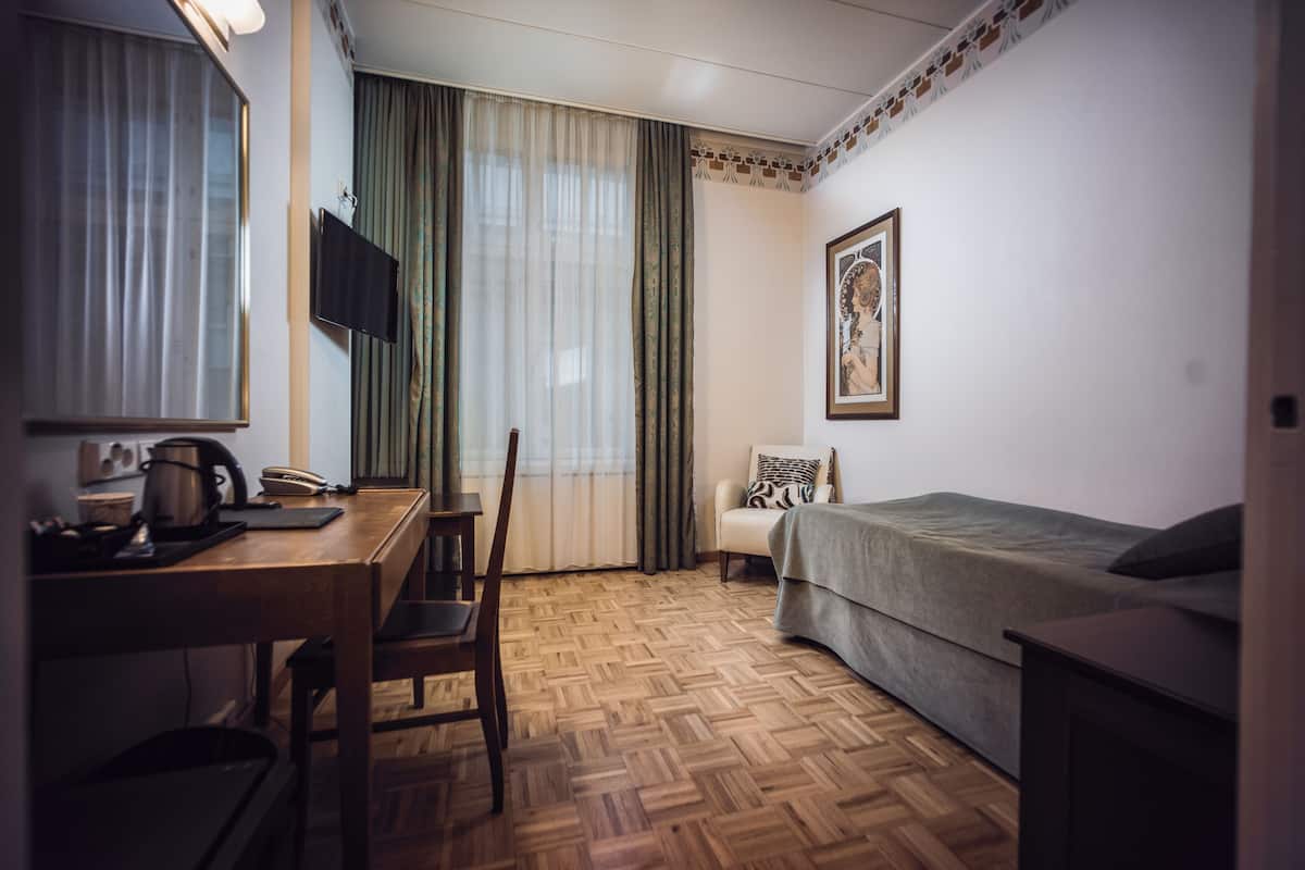 Hotellihuone, jossa on puinen kalanruotokuvioinen lattia, sänky, työpöytä tuolilla, seinälle asennettu televisio ja ikkuna, jossa on verhot.