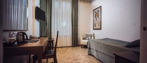 A hotel room with a wooden herringbone floor, a bed, a desk with a chair, a television mounted on the wall, and a window with curtains.