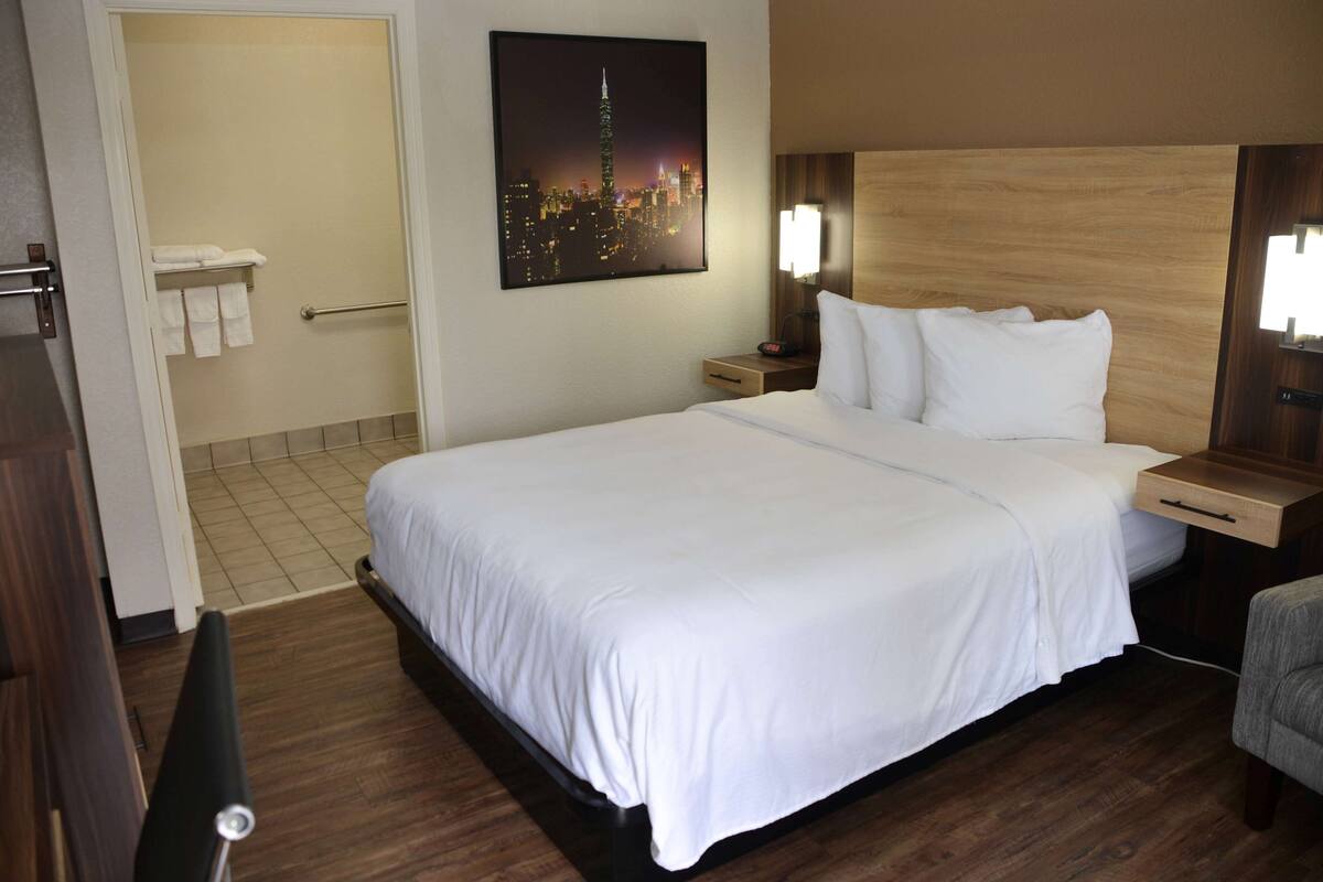 Une chambre d’hôtel avec un grand lit, une table de chevet, une lampe, une tête de lit en bois et un tableau représentant un paysage urbain accroché au mur.