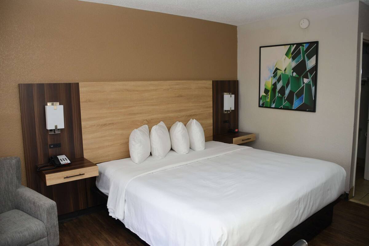 Une chambre d’hôtel avec un grand lit, une tête de lit en bois, deux tables de chevet avec des lampes, un fauteuil gris et une peinture abstraite moderne accrochée au mur.