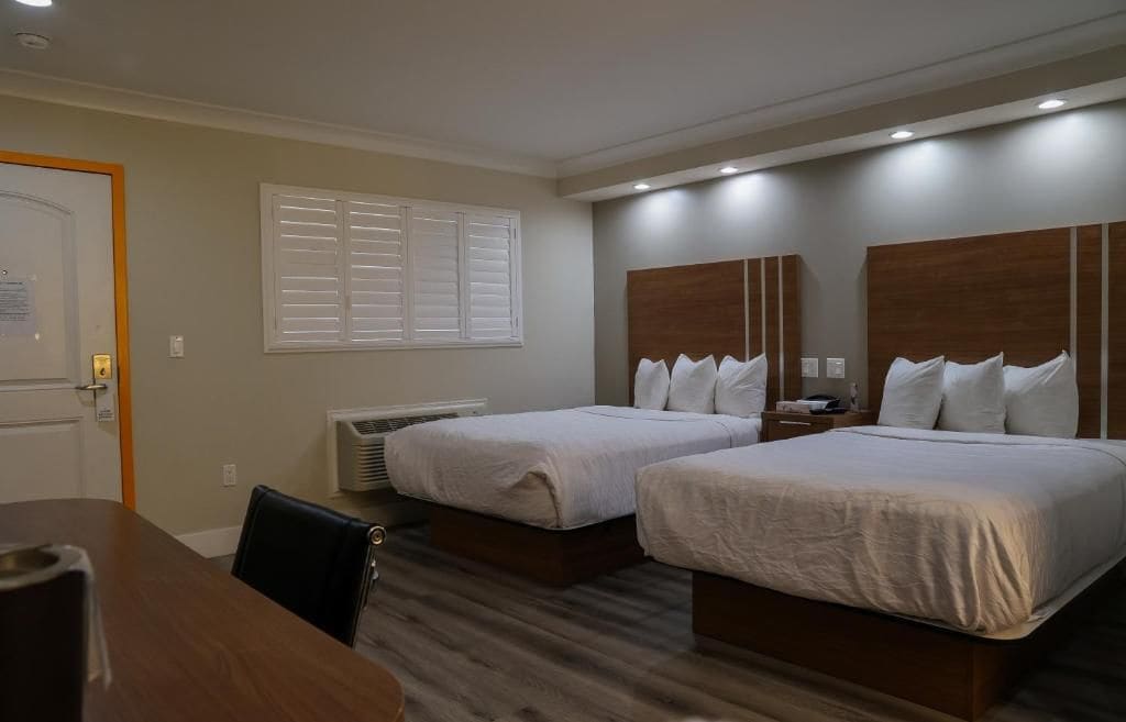 Une chambre d’hôtel avec deux lits, un bureau en bois, une chaise, une fenêtre avec des stores et un appareil de climatisation.
