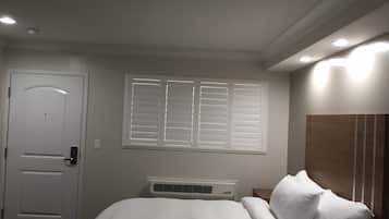Een slaapkamer met een bed, een witte deur, een raam met zonwering, een airconditioning en een nachtkastje met een lamp.