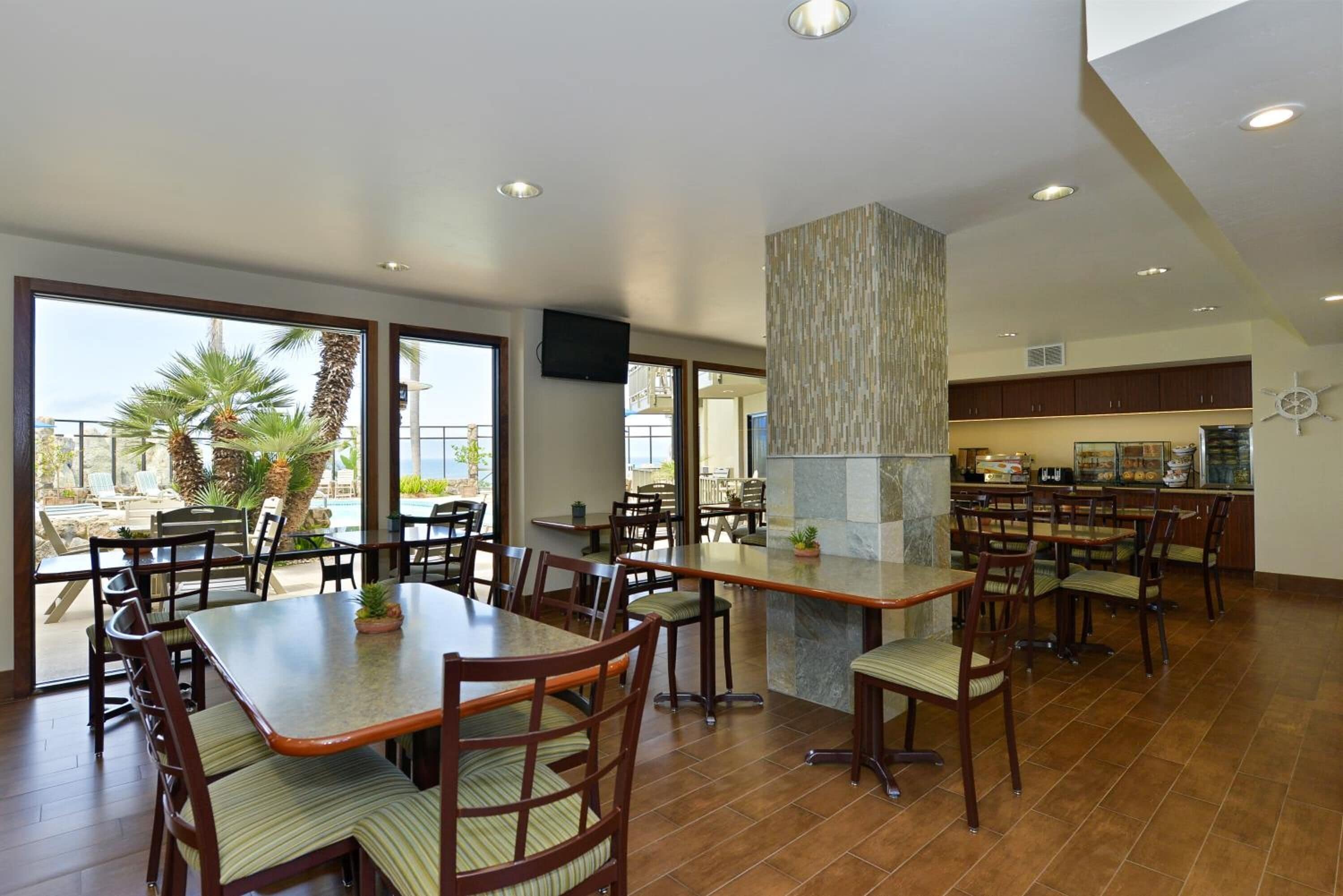 A dining area with multiple tables and chairs, a kitchen area with a buffet, and large windows offering a view of palm trees and a pool.