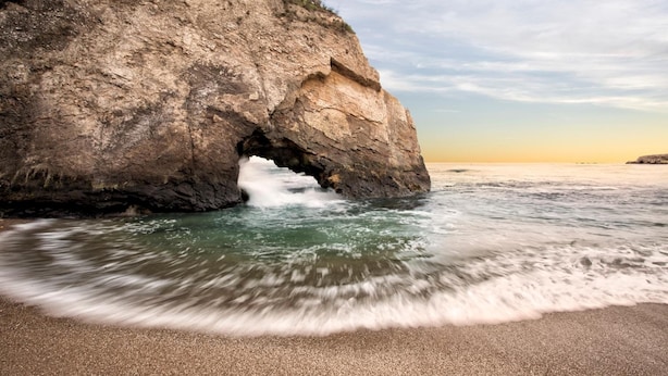 Un paisaje costero con un acantilado rocoso, olas rompiendo en una playa arenosa y una gaviota volando en el cielo.