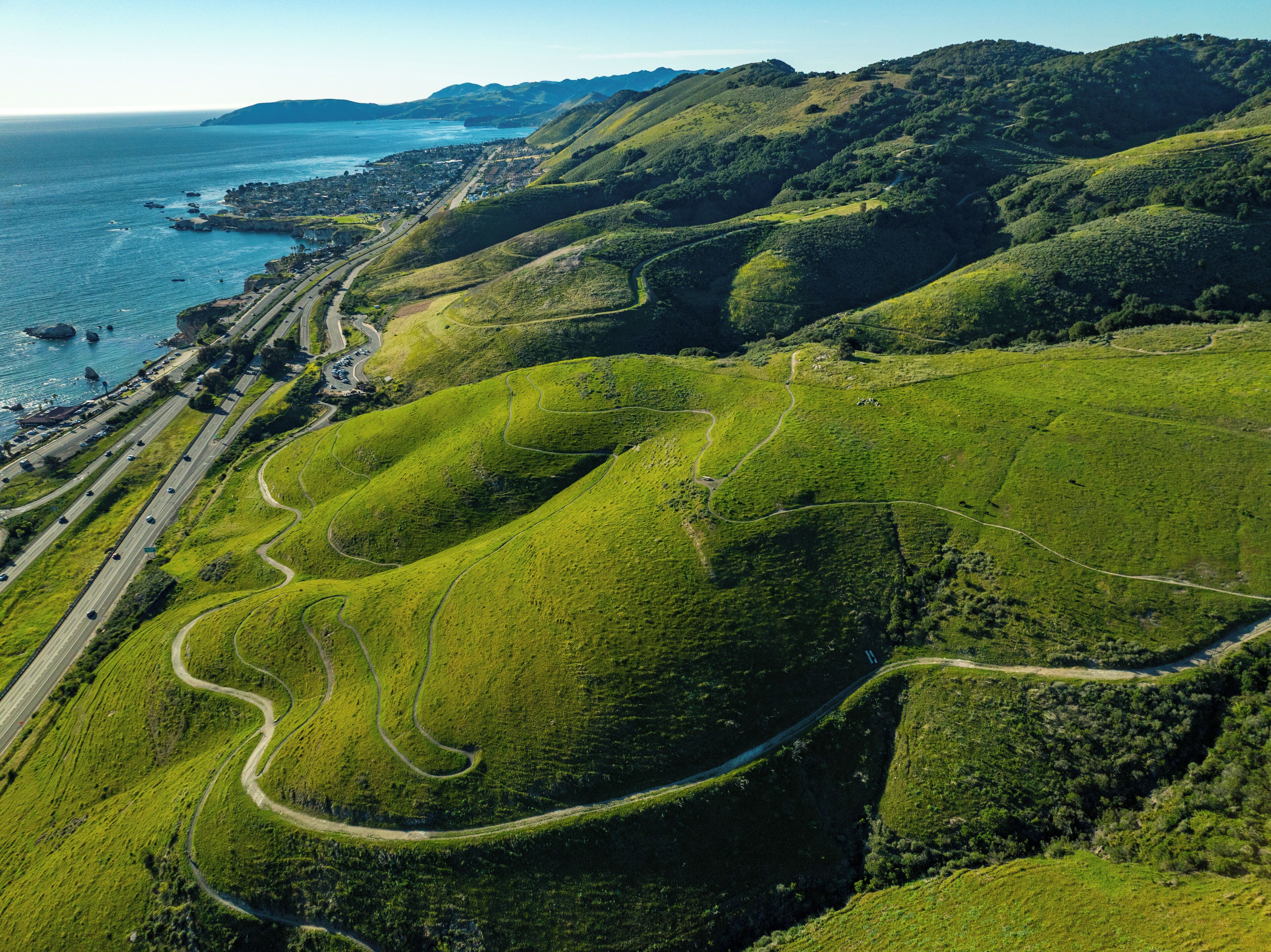 Vista aérea de un paisaje costero con carreteras sinuosas, colinas verdes y una masa de agua.