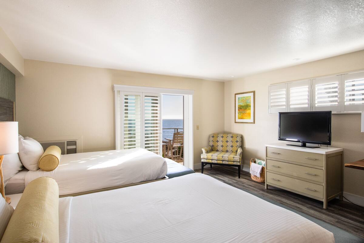 Habitación de hotel con dos camas, una televisión, una silla y vista al océano desde la ventana.