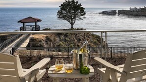 Una terraza con dos sillas, una mesa con dos copas de vino, una botella de champán y vistas al océano y a la costa rocosa.