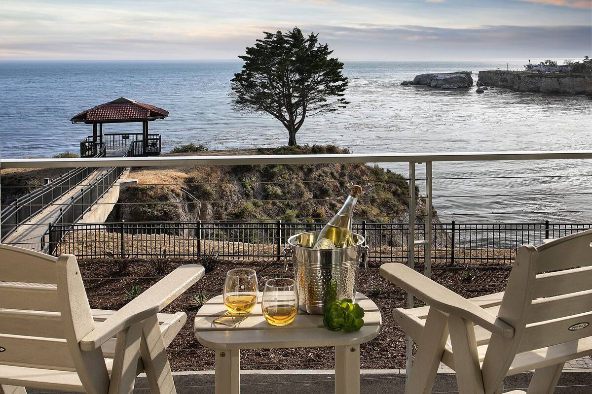 Una terraza con dos sillas, una mesa con dos copas de vino, una botella de champán y vistas al océano y a la costa rocosa.