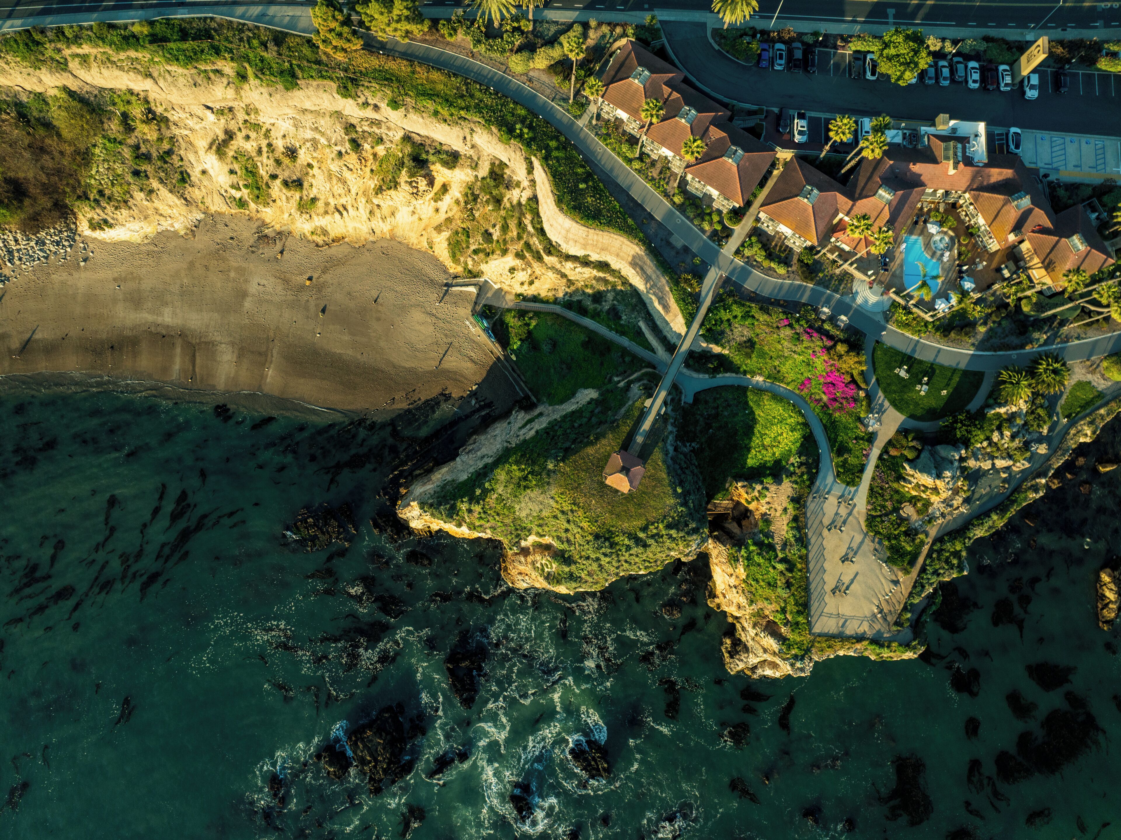 Vista aérea de una zona costera con playa, una carretera curva y un conjunto de edificios con tejados rojos.
