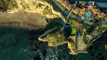 Vista aérea de una zona costera con playa, una carretera curva y un conjunto de edificios con tejados rojos.