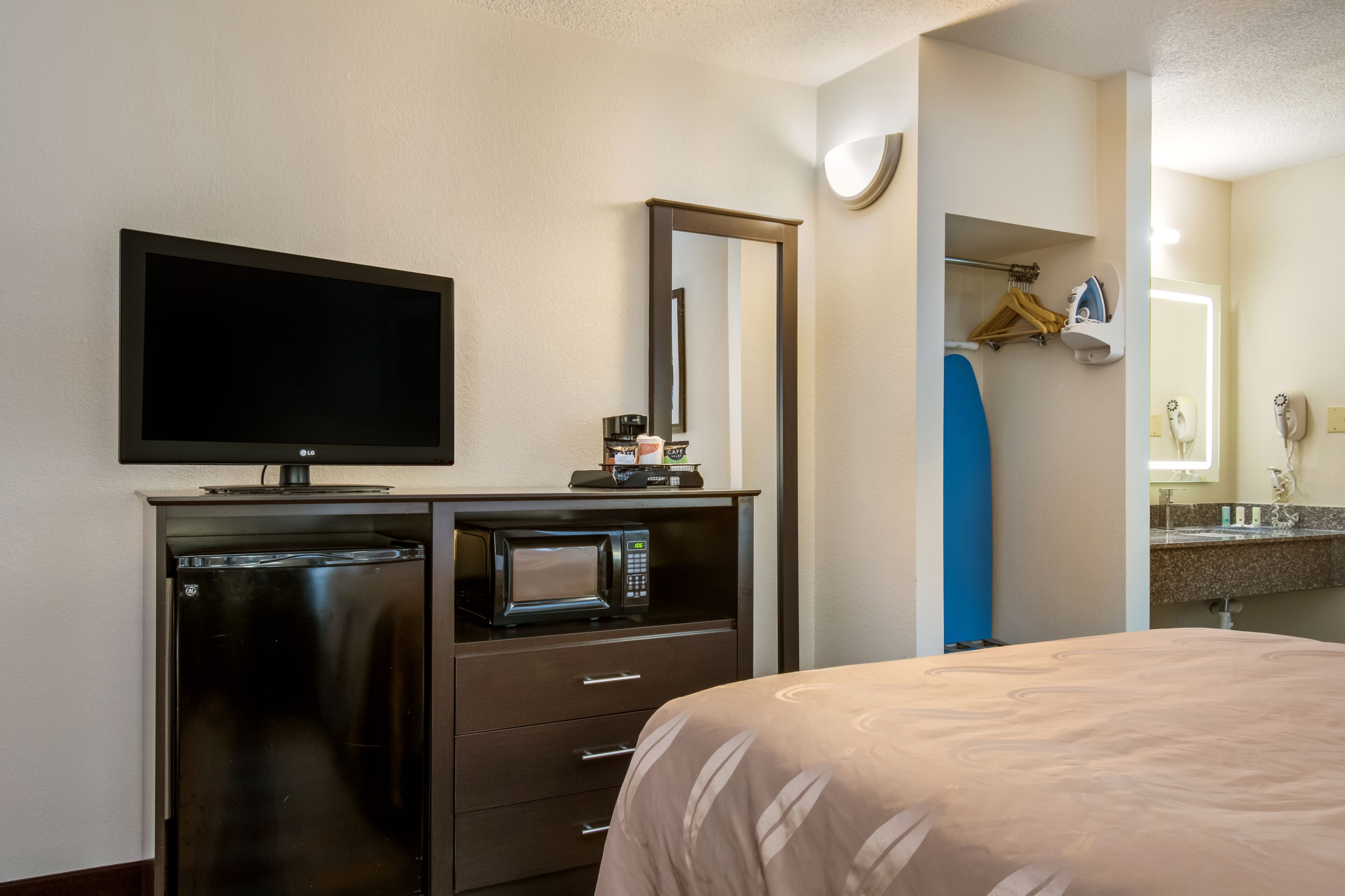 A hotel room with a flat-screen TV, a microwave, a mini-fridge, a bed, a bathroom with a sink, and a mirror.