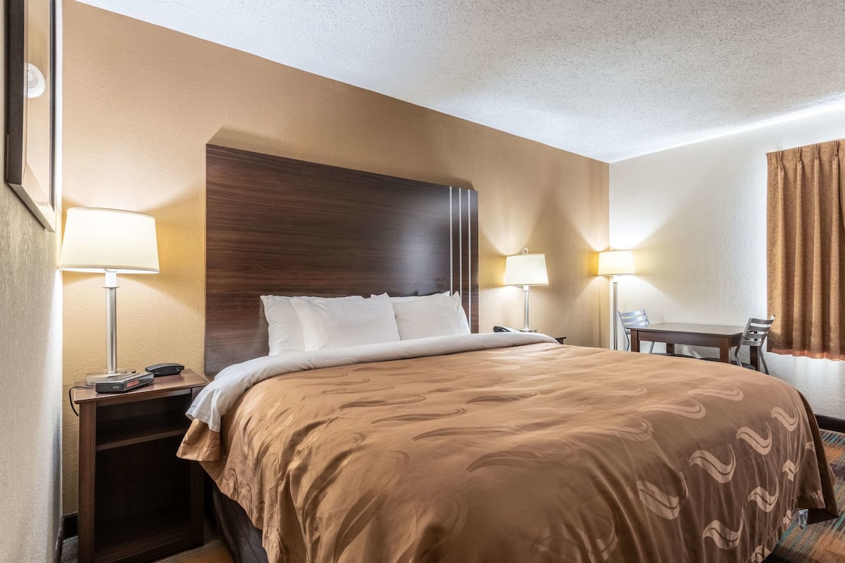 A hotel room with a large bed, two bedside tables with lamps, a wooden headboard, and a small table with chairs.