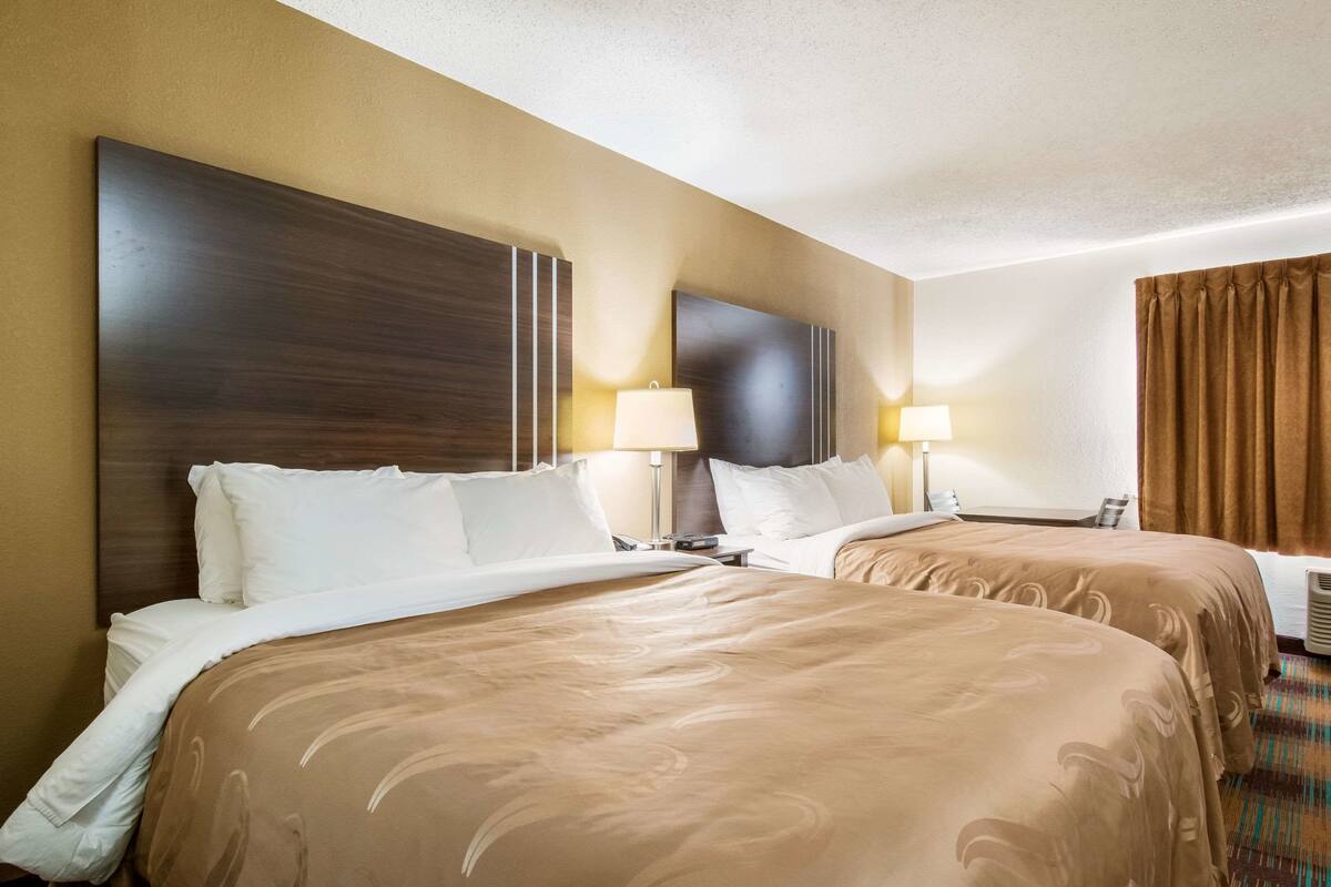 A hotel room with two beds, each with a headboard, bedside tables with lamps, and a large window with curtains.