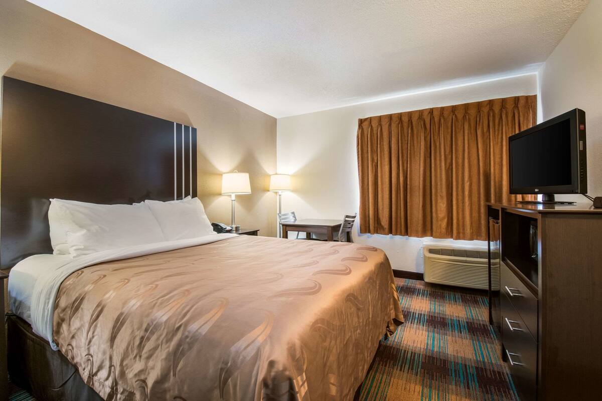 A hotel room with a large bed, a desk, two lamps, a television, and a window with curtains.