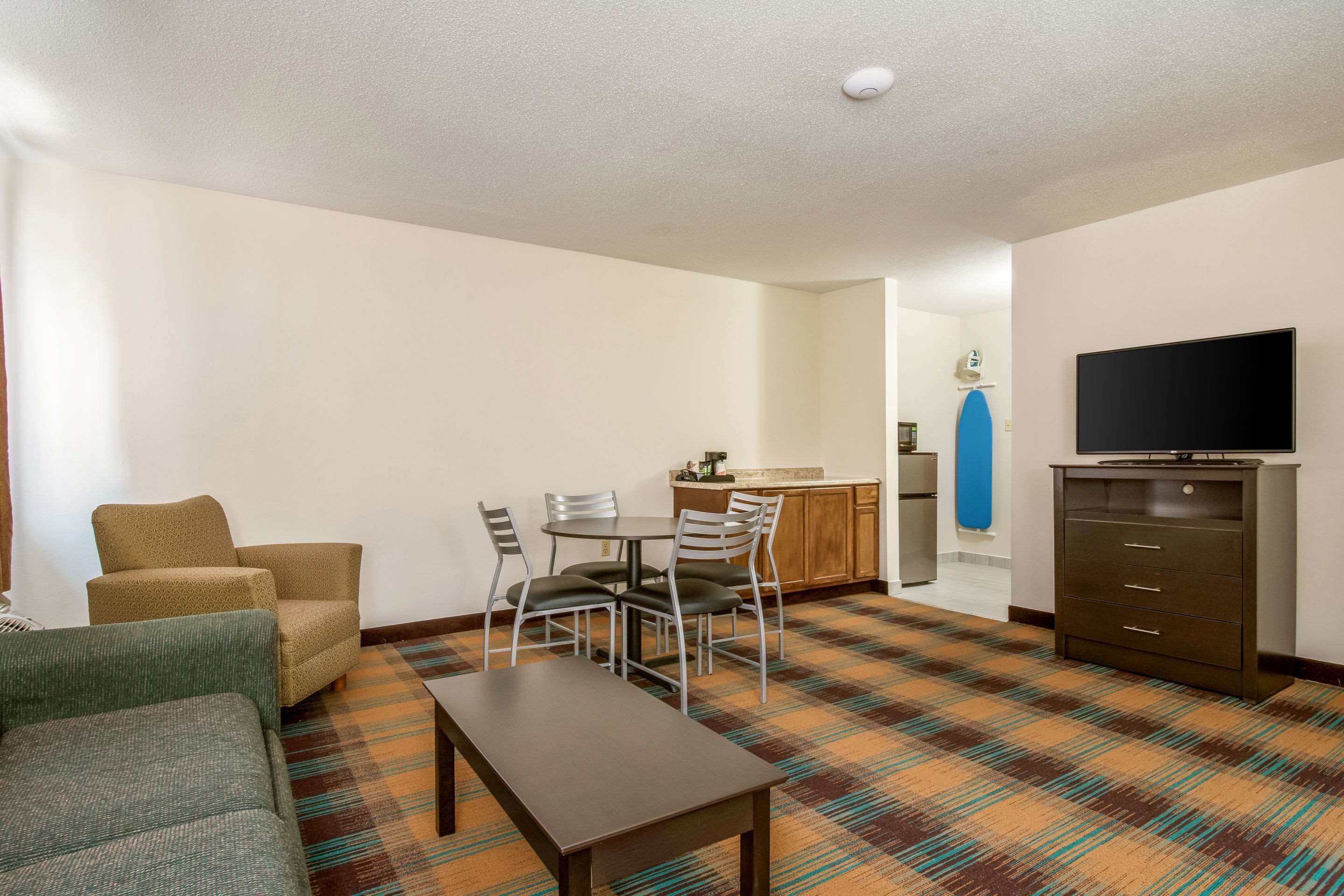A hotel room with a sofa, a dining table with chairs, a kitchen area, and a television on a cabinet.