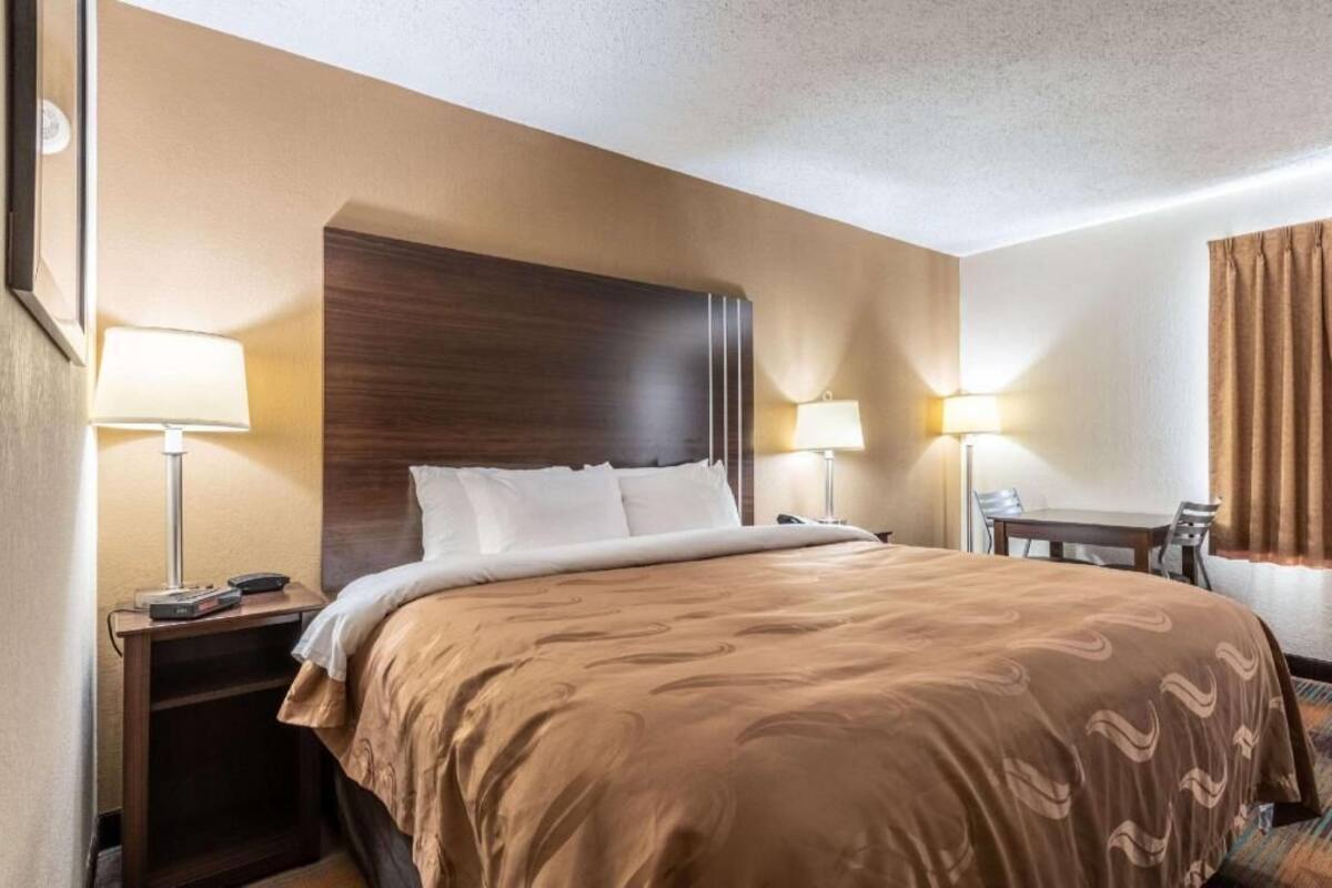 A hotel room with a large bed, two bedside lamps, a wooden headboard, a small table with chairs, and a curtain on the window.