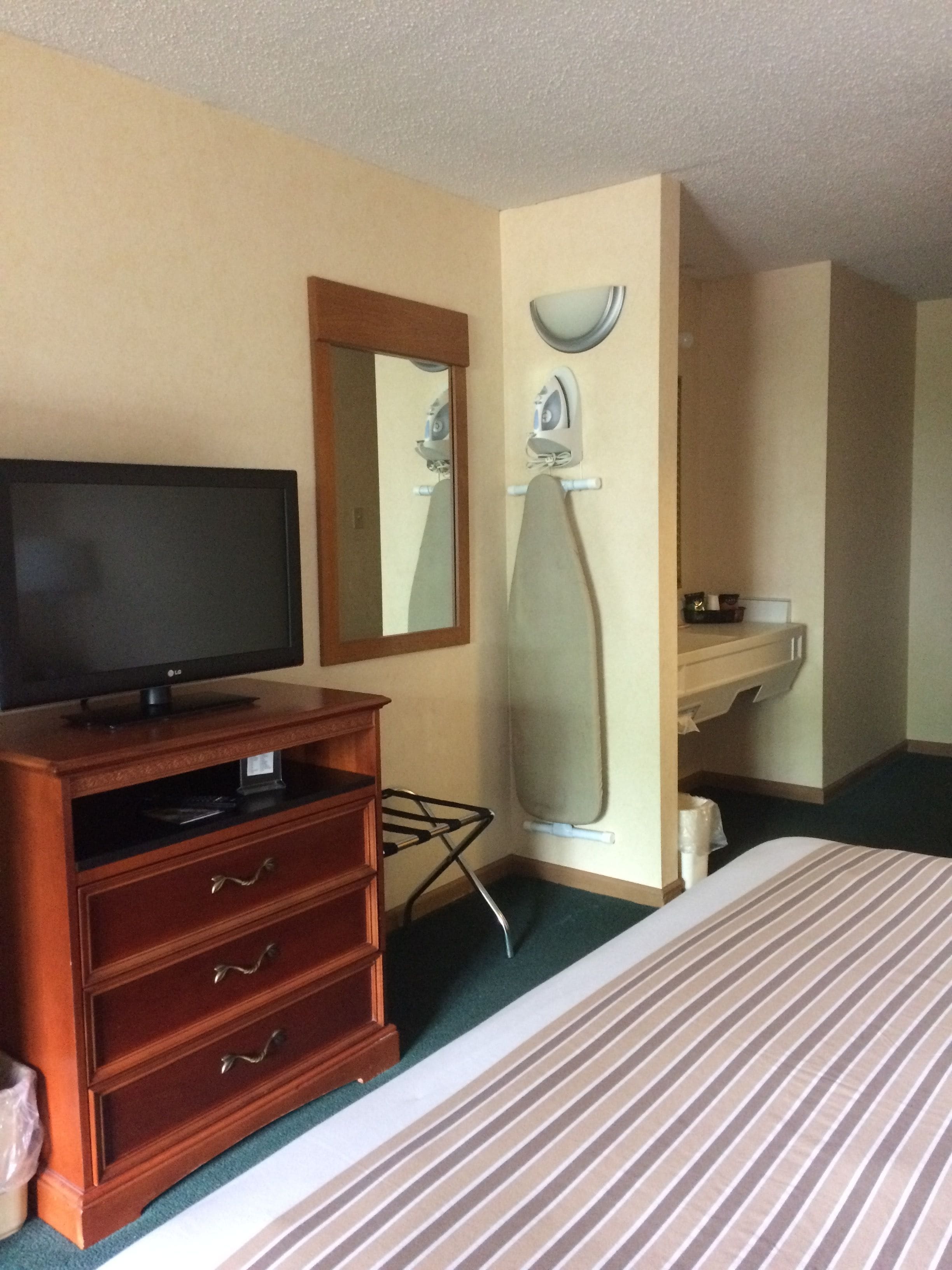 A hotel room with a bed, a television on a wooden dresser, a mirror, an ironing board, and a sink.