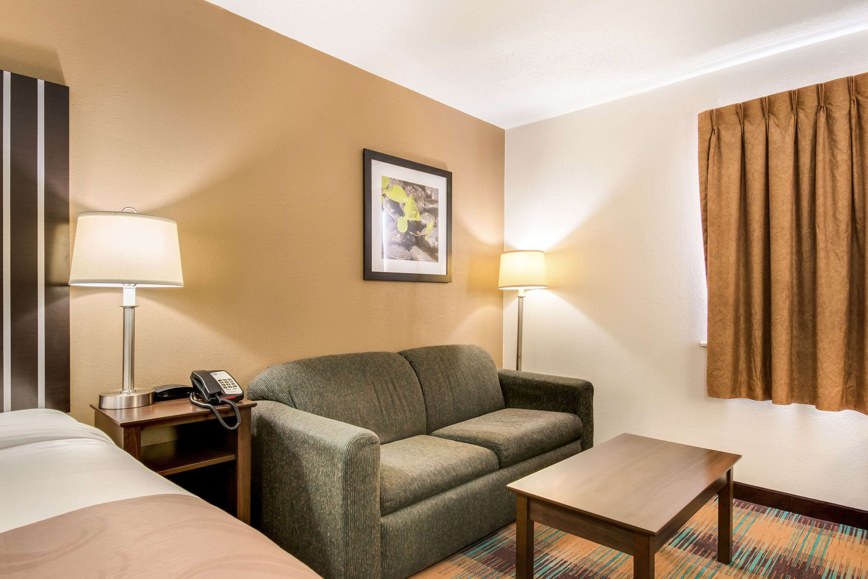 A hotel room with a bed, a sofa, a small table, two bedside lamps, a telephone on the nightstand, and a framed picture on the wall.