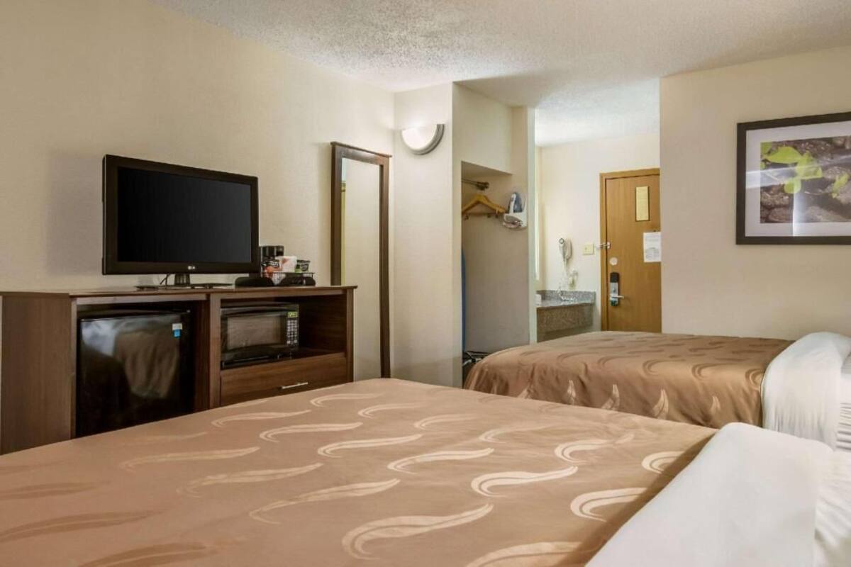 A hotel room with two beds, a television, a mini-fridge, a mirror, and a door leading to a bathroom.