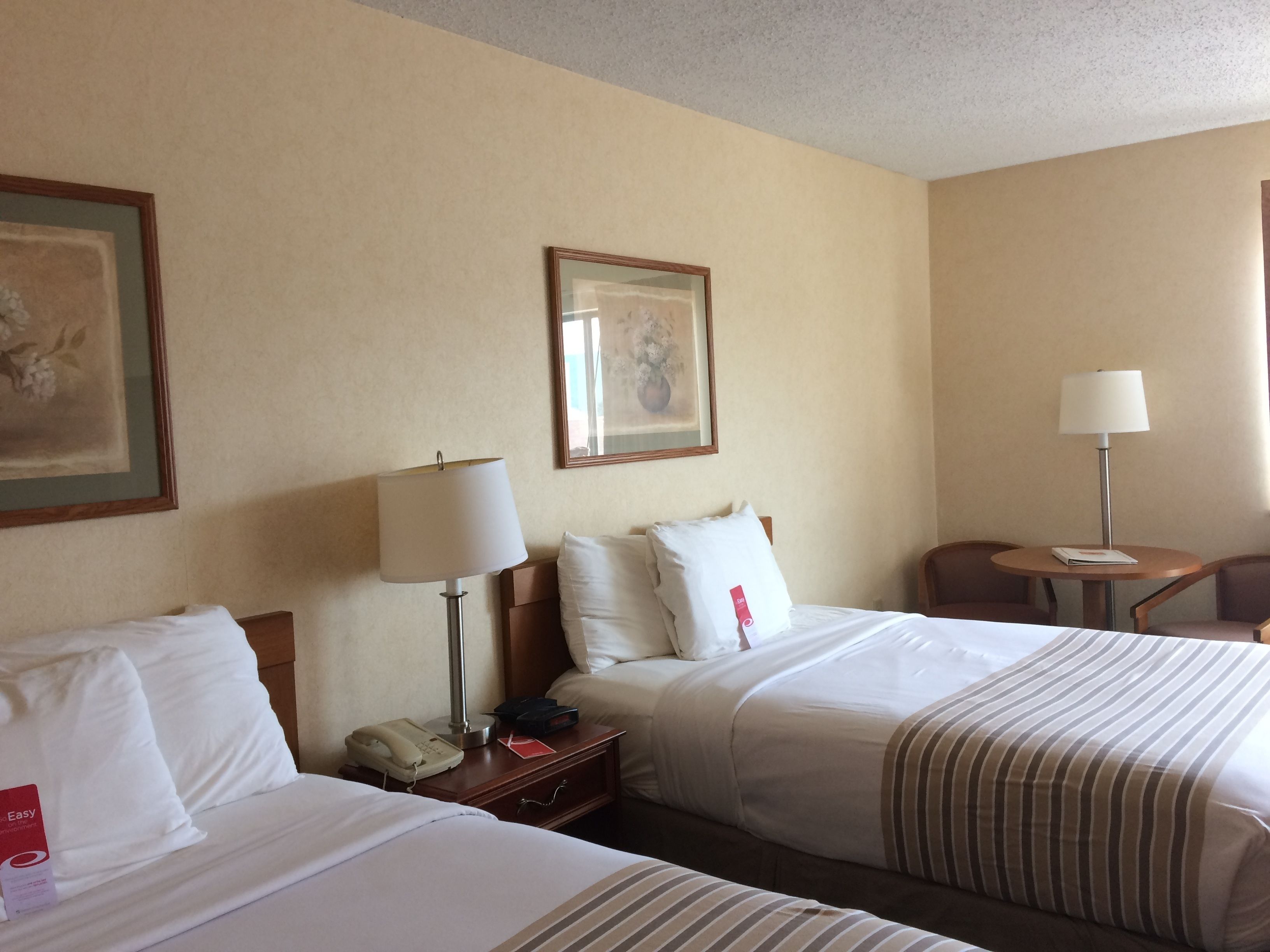 A hotel room with two beds, a bedside table with a lamp, a chair, and a framed picture on the wall.
