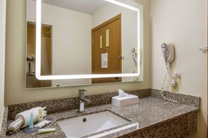 A bathroom with a granite countertop, a white sink, a mirror with a light, a towel rack, a hairdryer, and a phone charger.