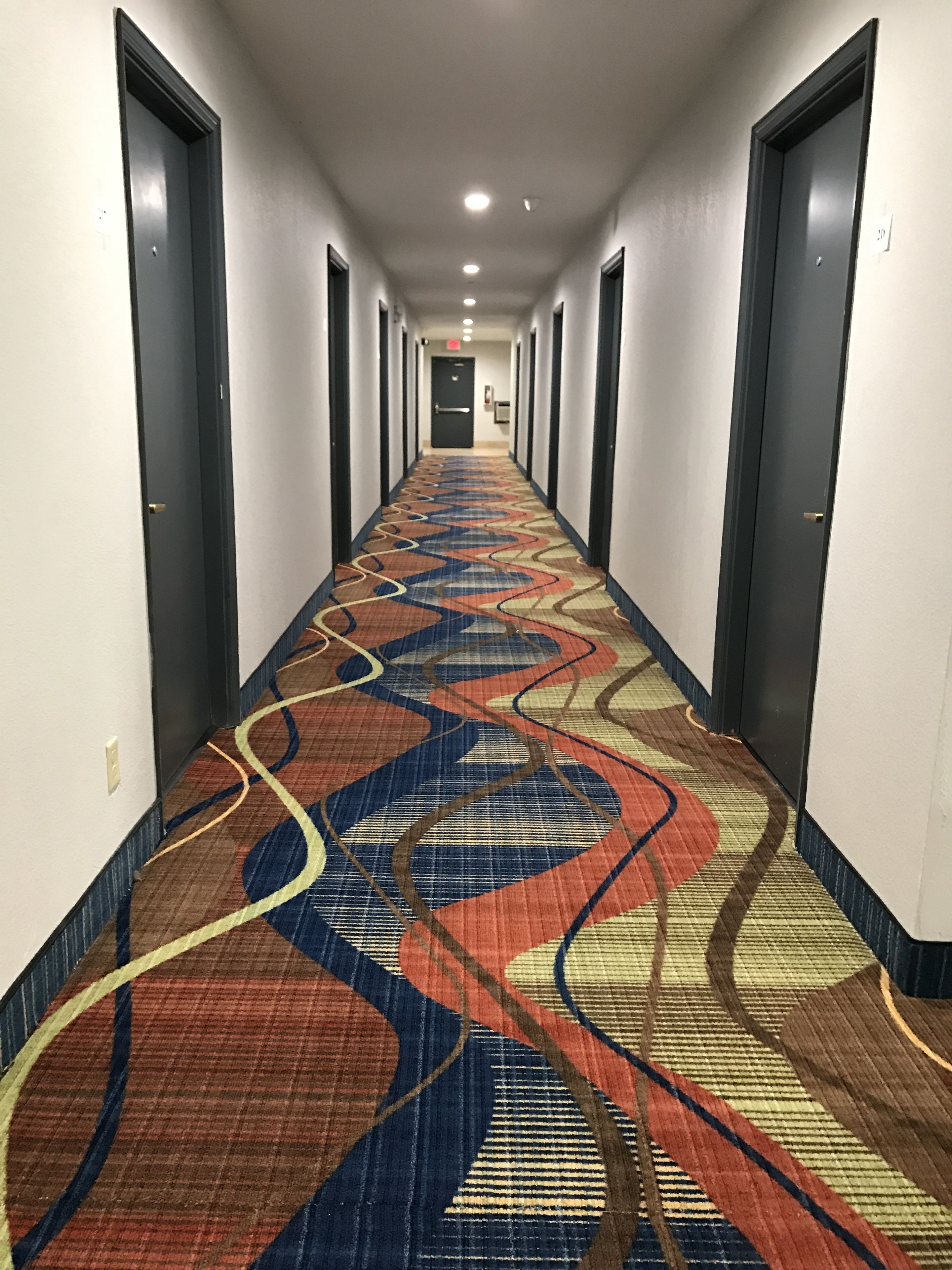 A hotel hallway with a patterned carpet, doors on both sides, and a light fixture on the ceiling.