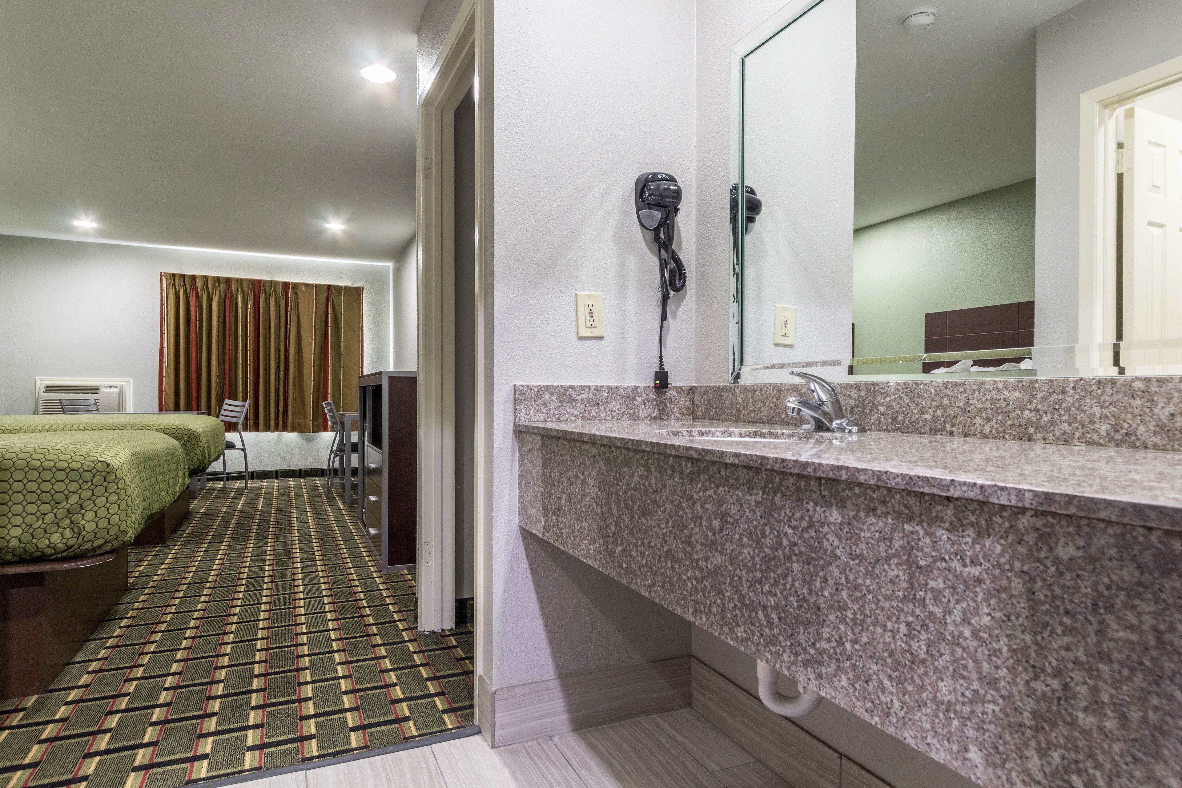 A hotel room with a bathroom featuring a granite countertop sink, a mirror, and two hair dryers on the wall. There is a bed with green bedding and a patterned rug on the floor.