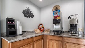 A kitchen with wooden cabinets, a coffee machine, a juice dispenser, and a shelf with cups and a decorative item. - Scottish Inn and Suites (Tomball)