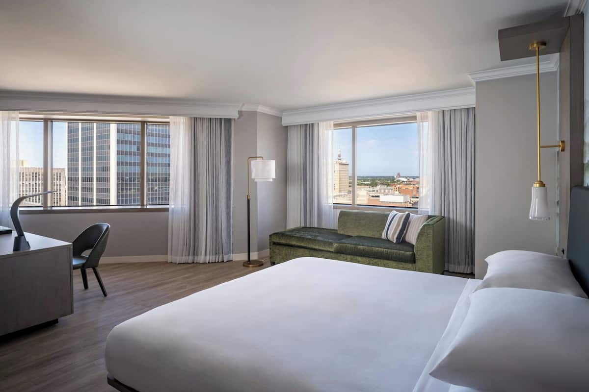 A hotel room with a large bed, a desk, a chair, a floor lamp, and a view of the cityscape.