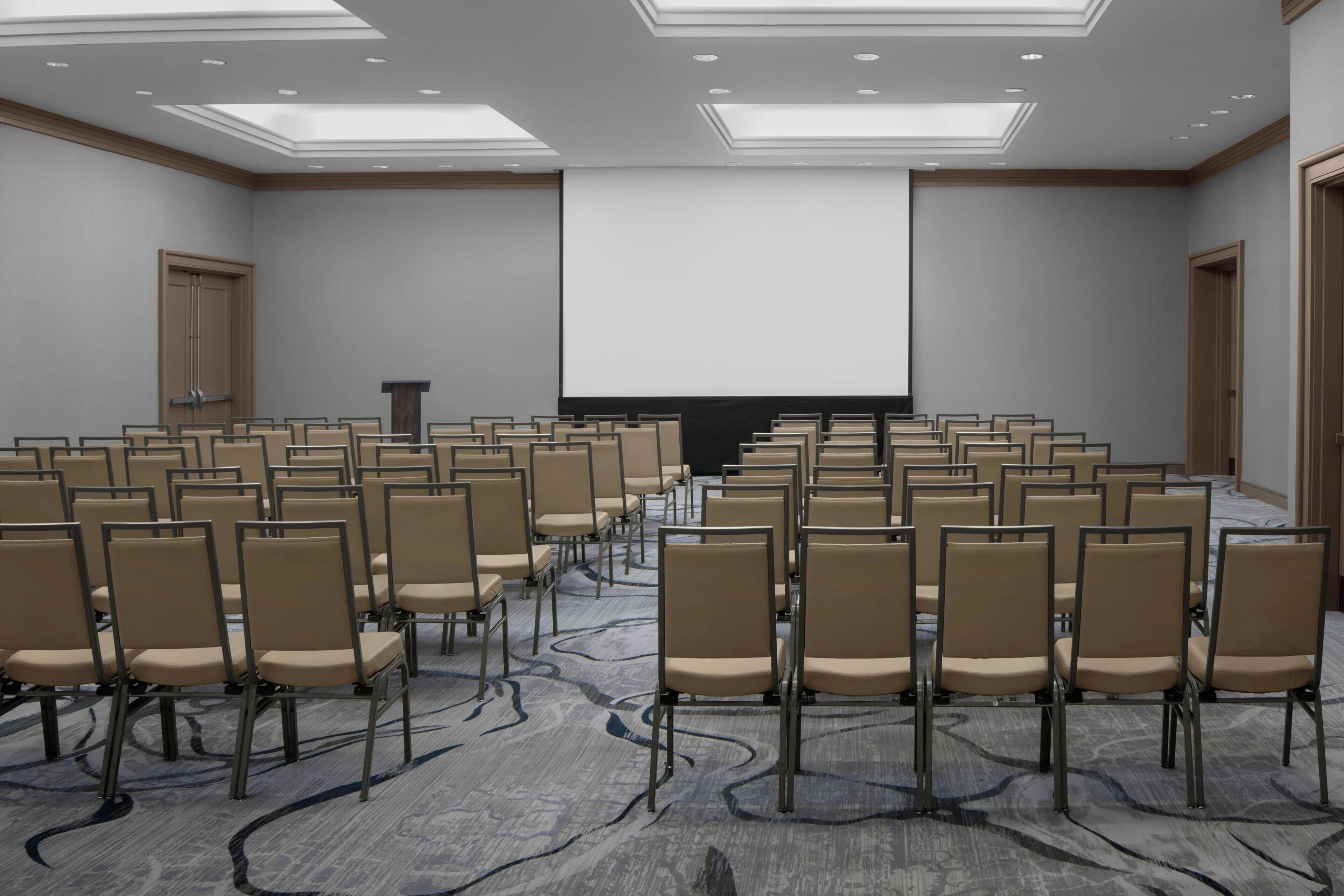 Une salle de conférence avec des rangées de chaises, un grand écran et un pupitre.