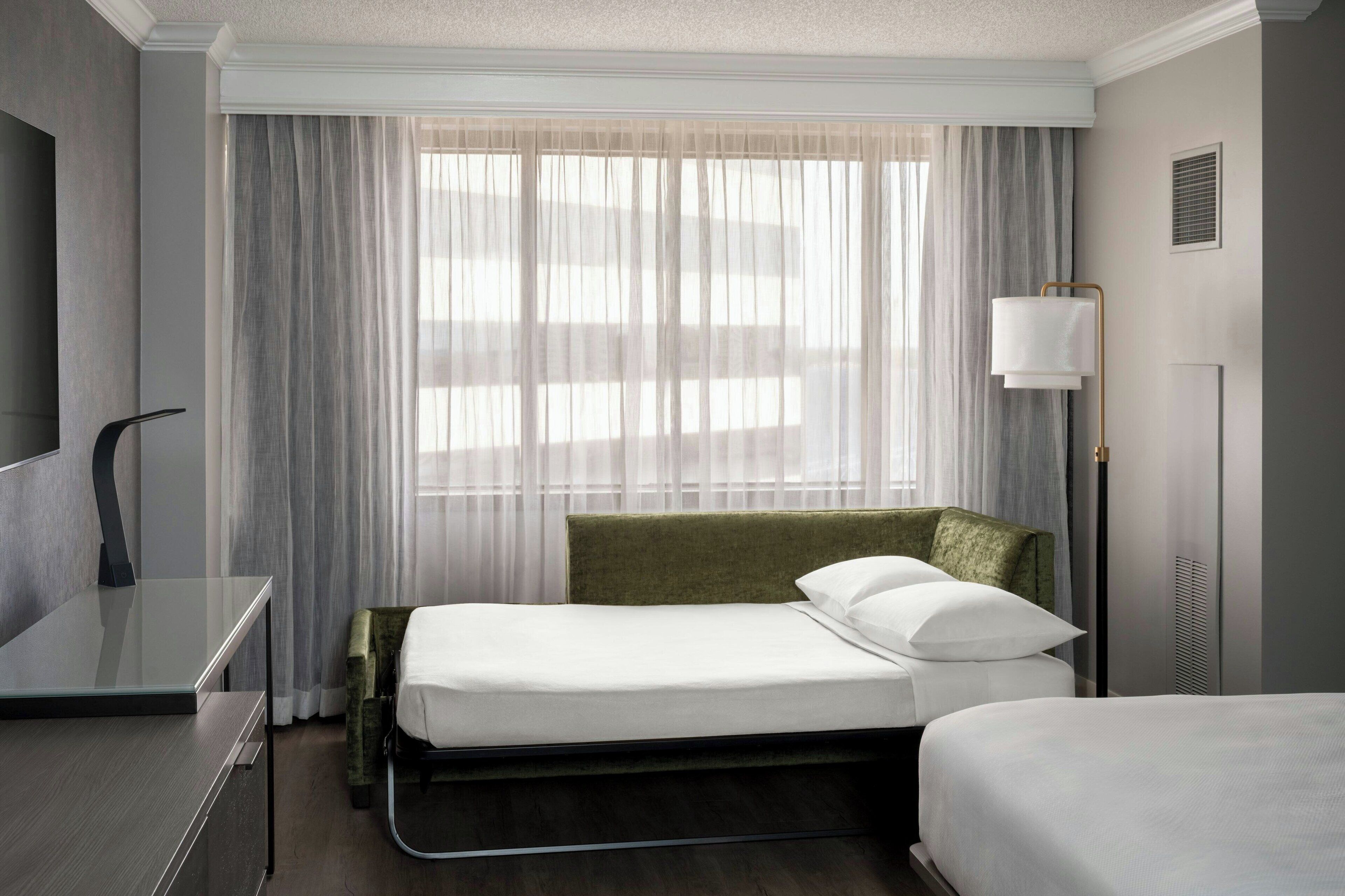 A hotel room with a bed, a desk, a lamp, and a window with curtains.
