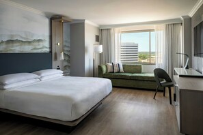 A hotel room with a large bed, a green sofa, a desk, and a view of the city through the window.