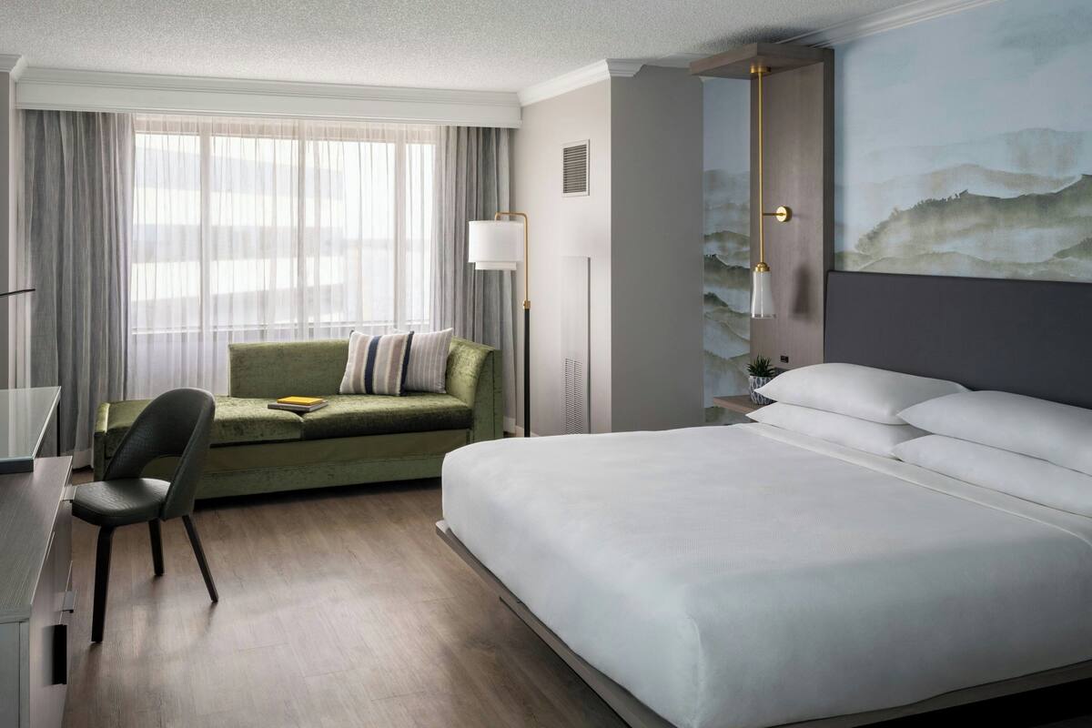 A hotel room with a bed, a desk, a chair, a sofa, and a large window with curtains.